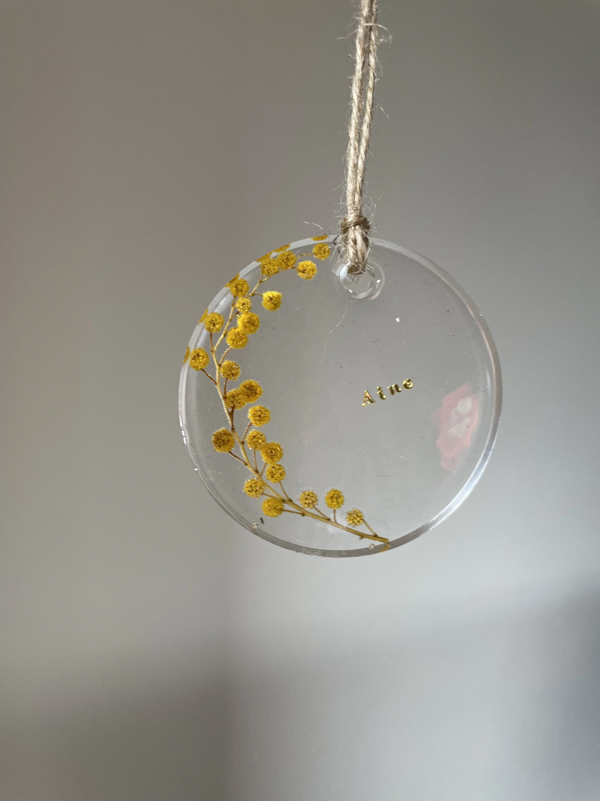 A round, transparent ornament with a dried yellow flower flower design and the word 'Aine' written inside, hanging from a piece of string.
