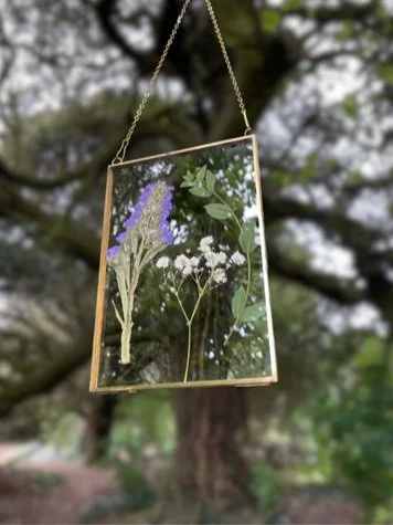 Hanging glass frame with pressed flowers and greenery outdoors.