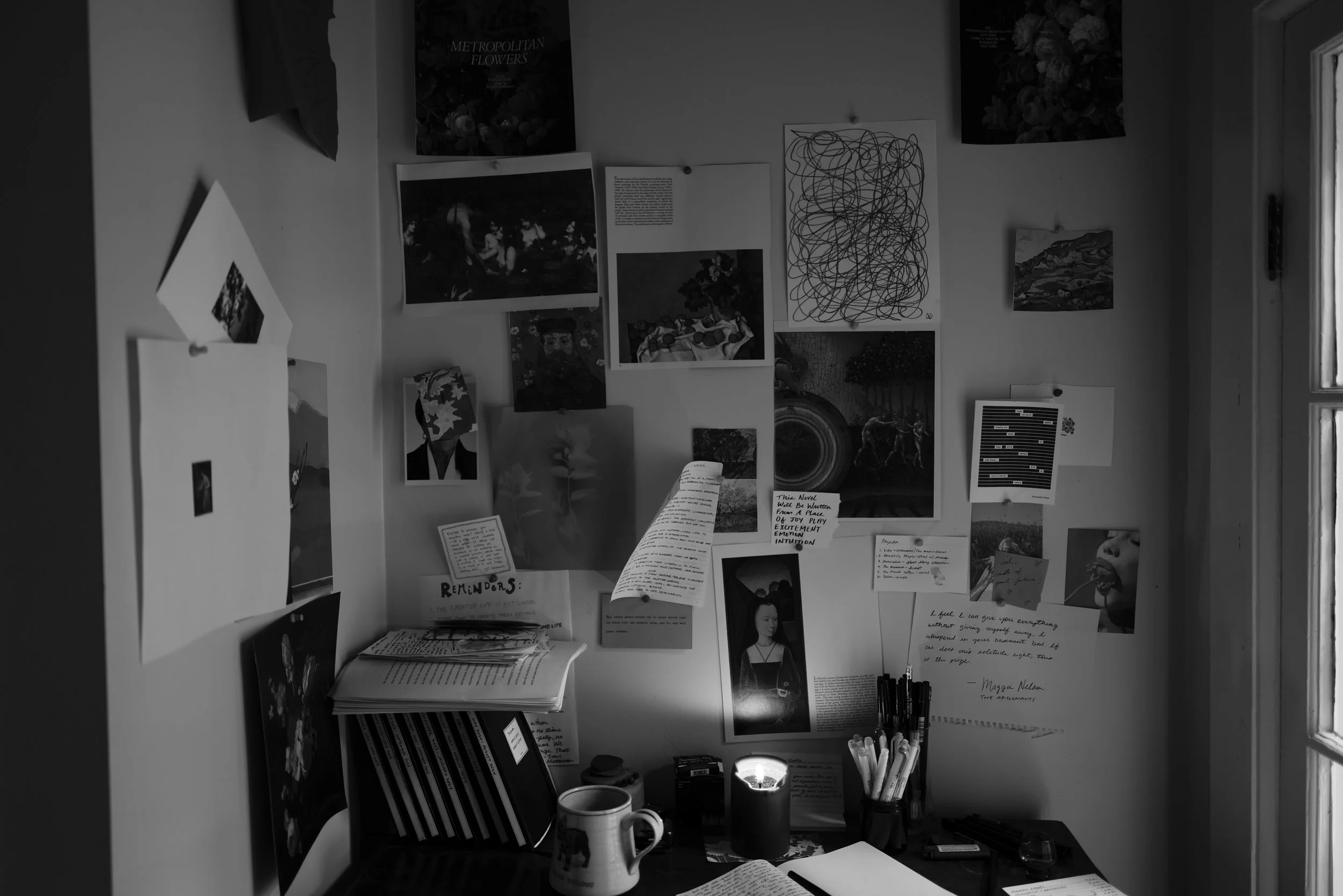 Black and white photograph of a creative workspace. There is a wall above a desk covered with art prints, notes, postcards, and drawings. A stack of paper and stationery items are on the desk with a lit candle adding light.