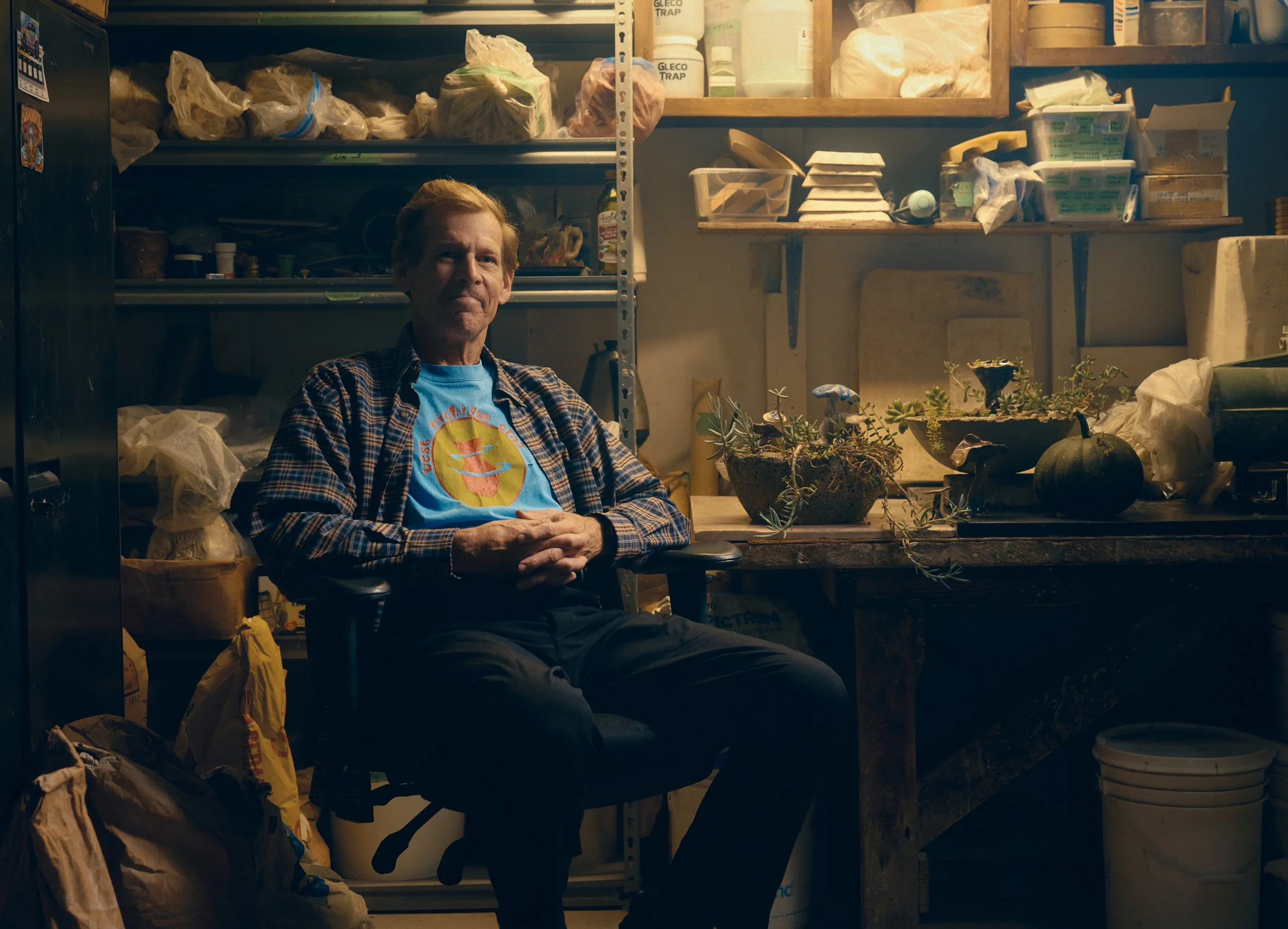 A wide-angle environmental commercial portrait of an artist in a cluttered workshop in Hamilton, MI