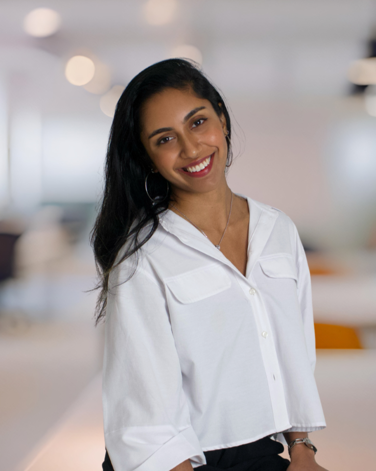 A woman with dark hair styled in loose waves, wearing a white button-up shirt, hoop earrings, a necklace, and smiling against a plain white background.