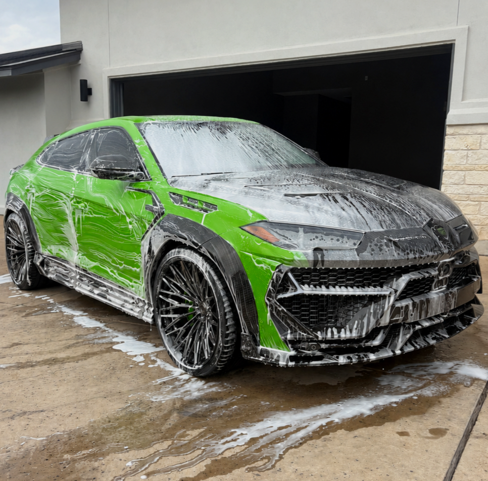 A green sports car being washed with soap and water, soap suds visible on the car's surface and on the ground in front of it.