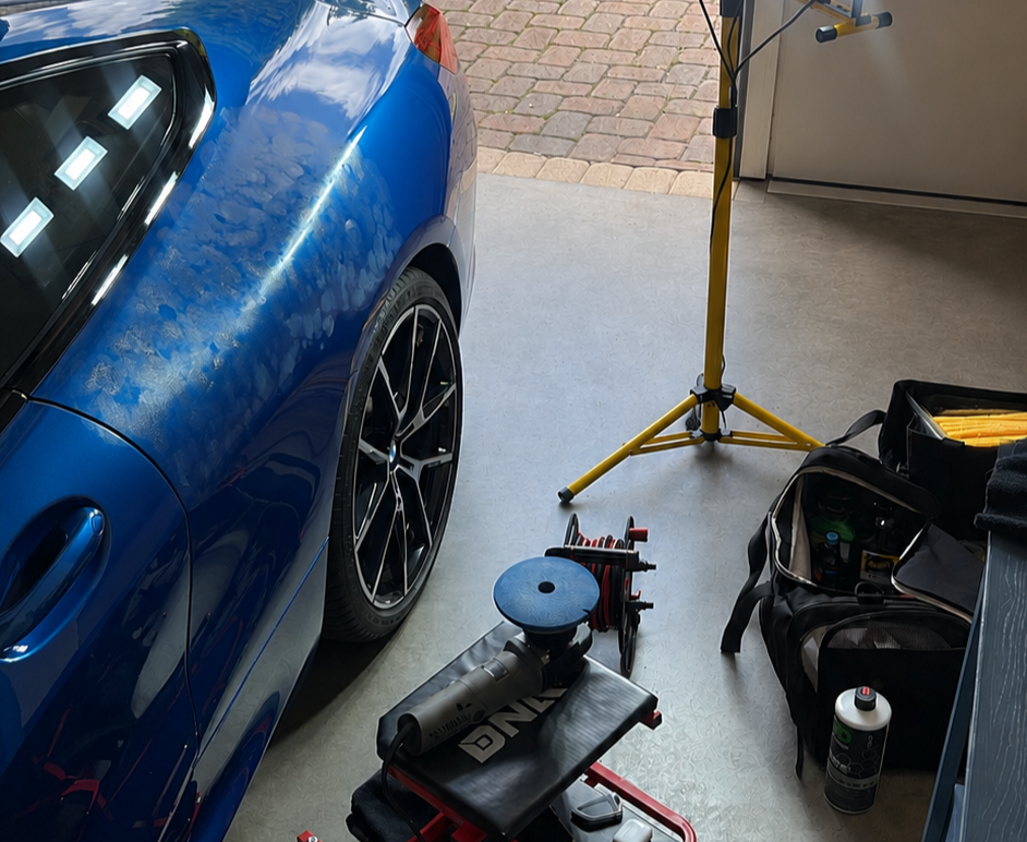 A blue sports car parked in a garage with tools and equipment nearby, including a yellow tripod, a black bag with supplies, and a polishing or detailing tool with a red cord.
