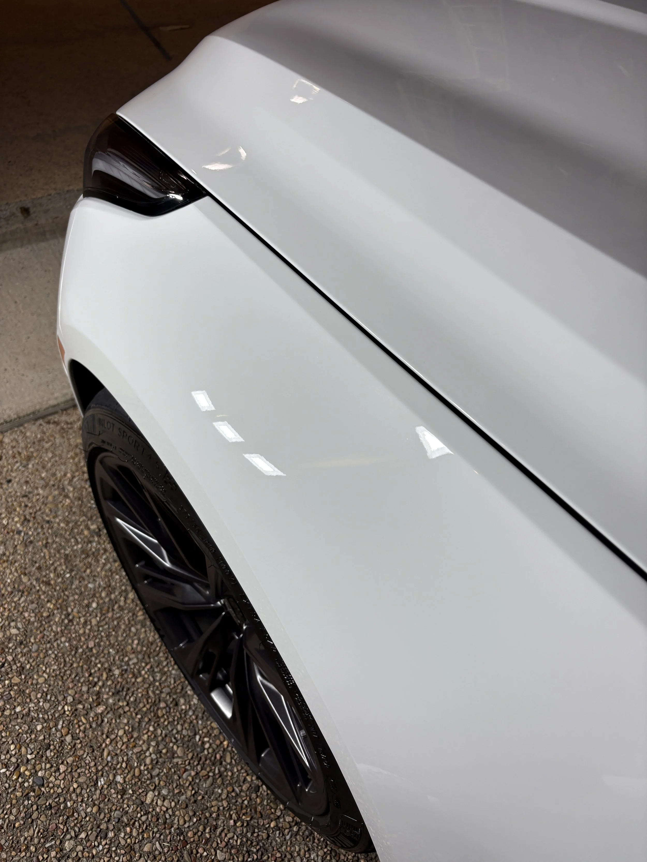 Close-up of the front right side of a white modern car, showing the headlight, part of the wheel, and the hood, parked on a gravel surface.