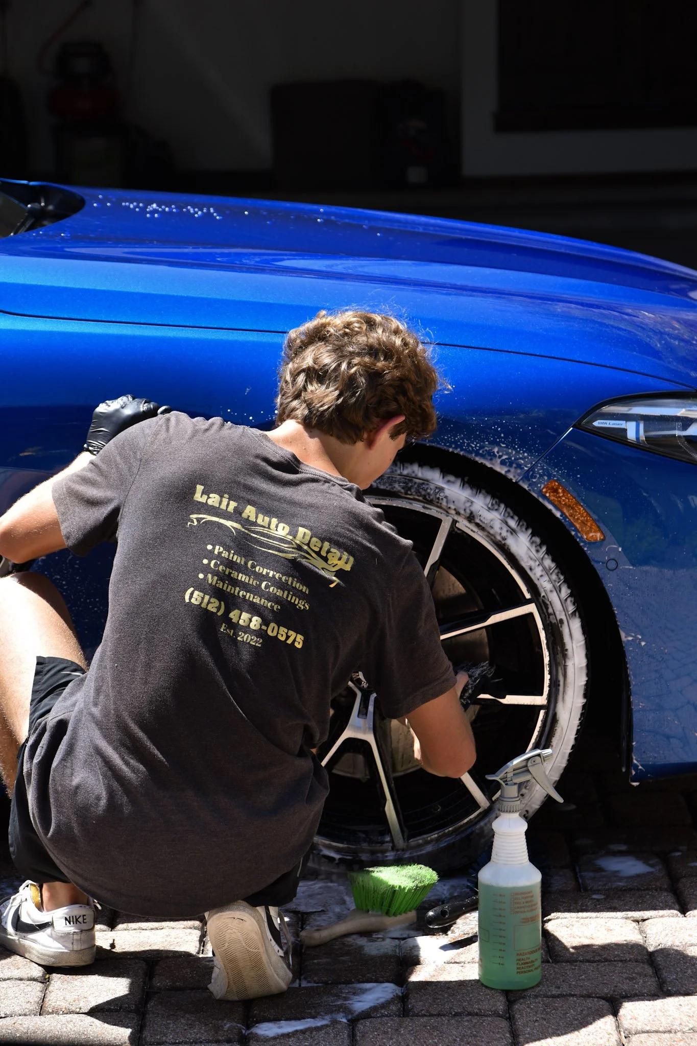 A person cleaning the wheels of a blue car during a car wash, with cleaning supplies on the ground.