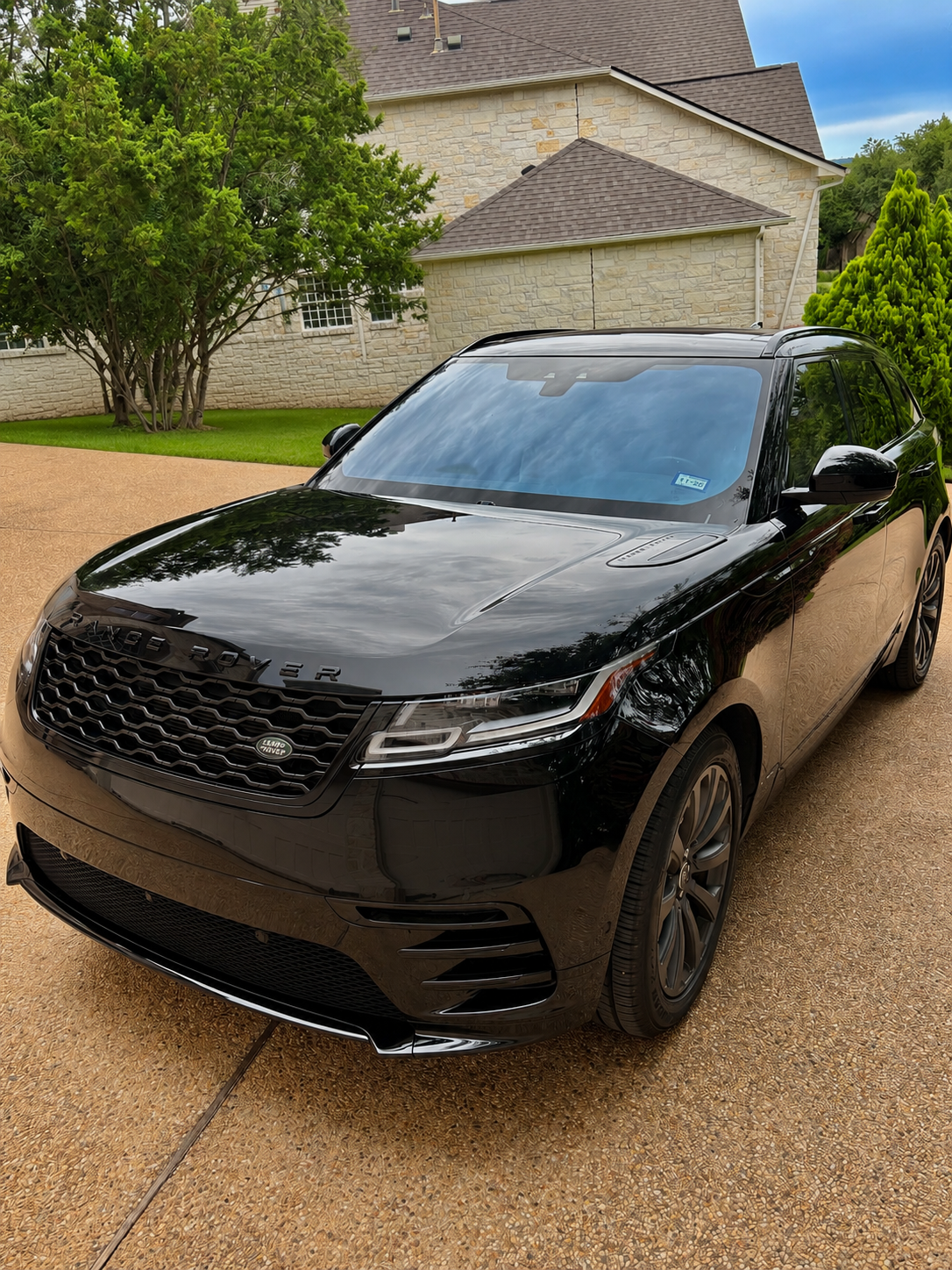 A black Land Rover SUV parked on a gravel driveway in front of a brick house with a sloped roof, surrounded by green trees and bushes.
