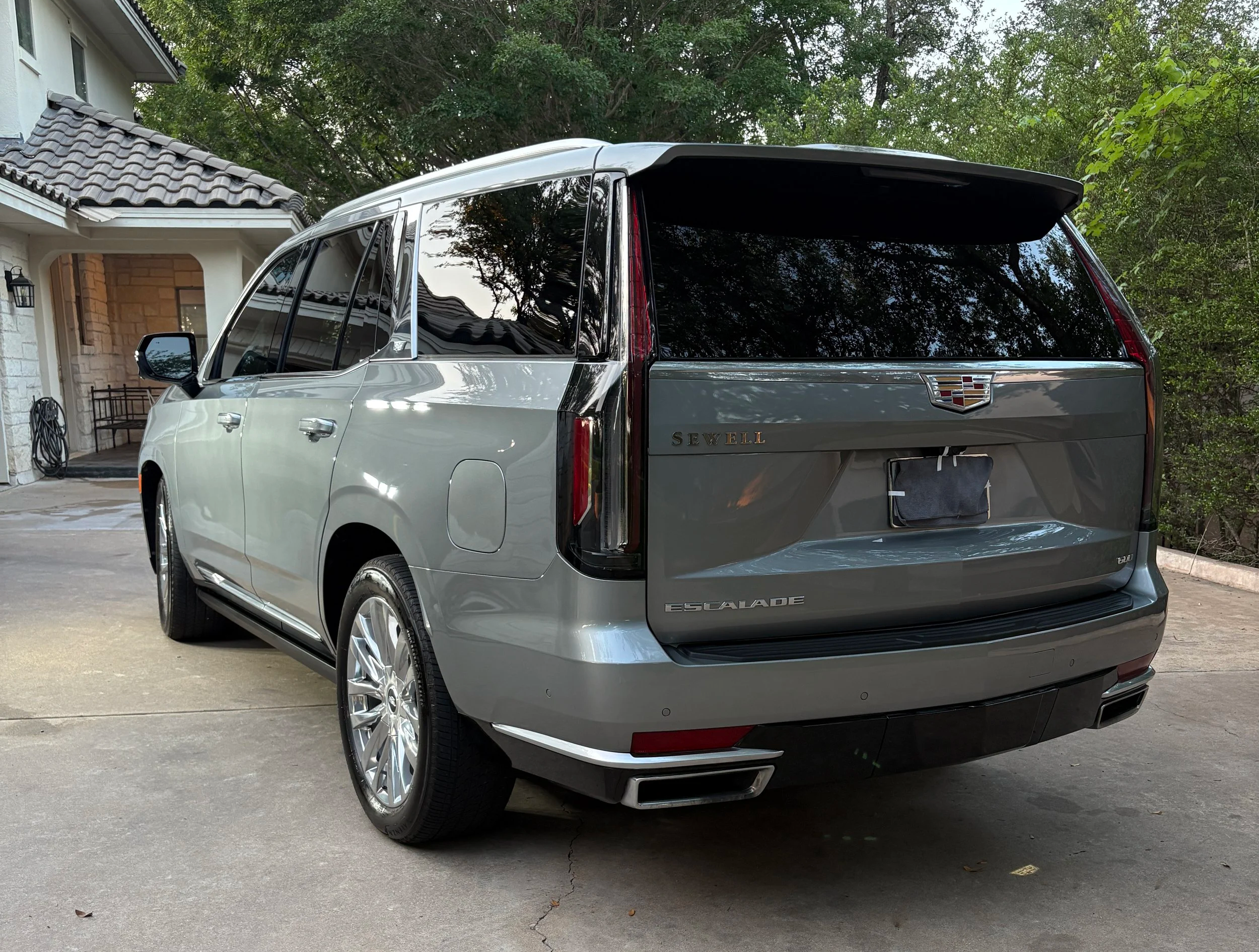 A silver Cadillac Escalade parked on a driveway near a house with trees in the background.