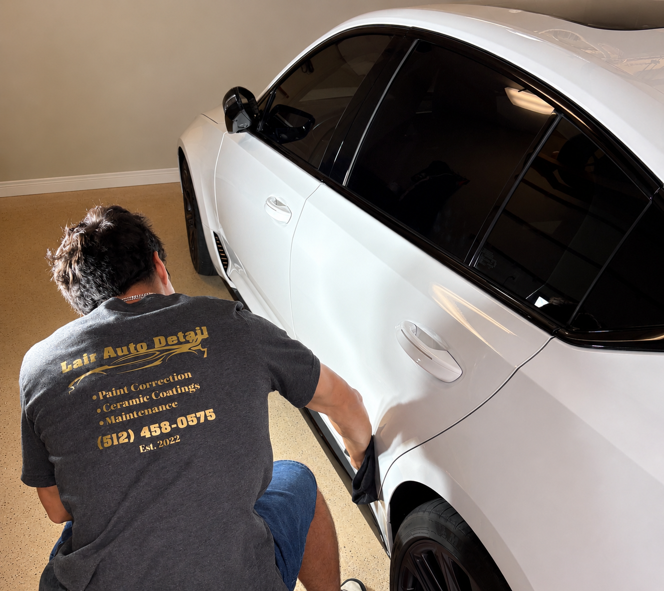 A person wearing a gray shirt with gold text and black pants is working on a white car inside a garage. The shirt has the words "Lair Auto Detail" and services like paint correction, ceramic coatings, and maintenance, along with a phone number.