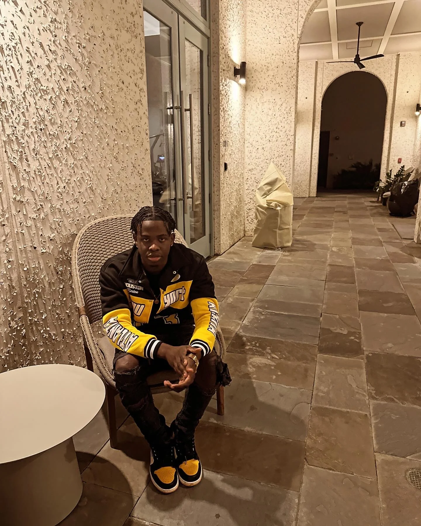 Person sitting on a chair in an outdoor corridor with textured walls, wearing a black and yellow jacket and matching sneakers, with a small round table nearby.