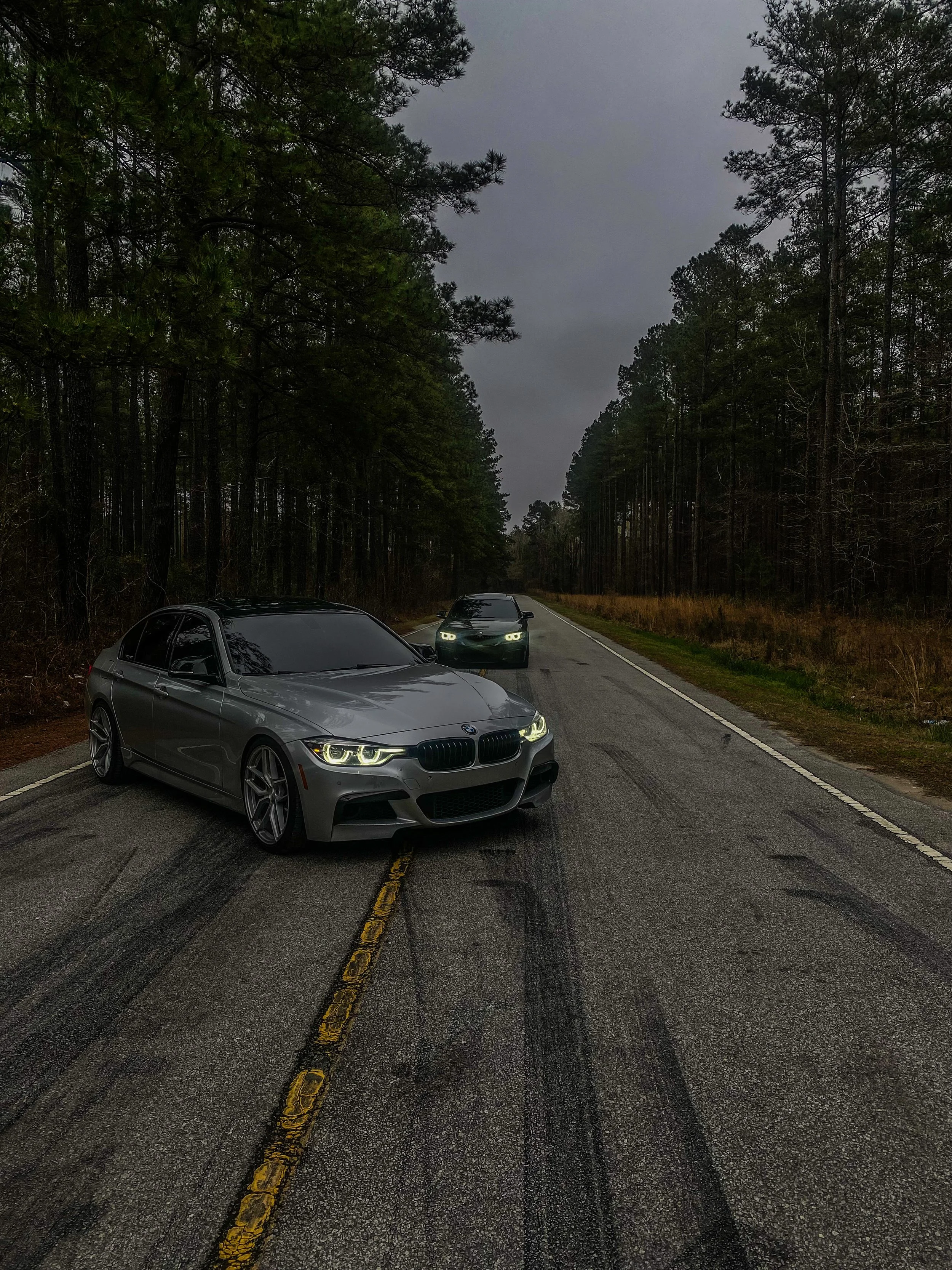 Two cars on a forest road with cloudy sky, one silver car in the foreground and another car further back.