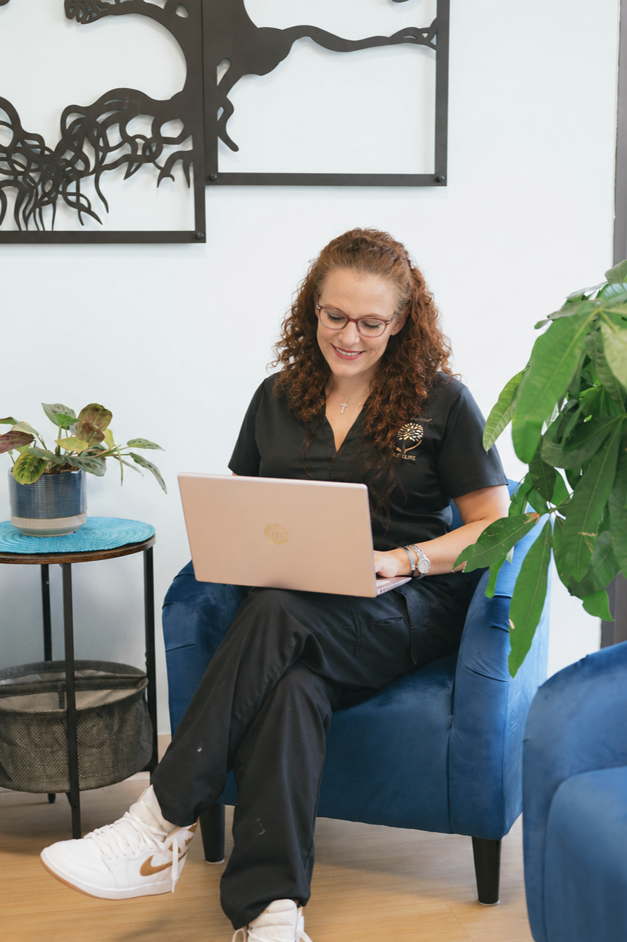 A woman with curly hair and glasses sitting in a blue armchair, working on a pink laptop in a modern, well-lit room with houseplants and abstract wall art.