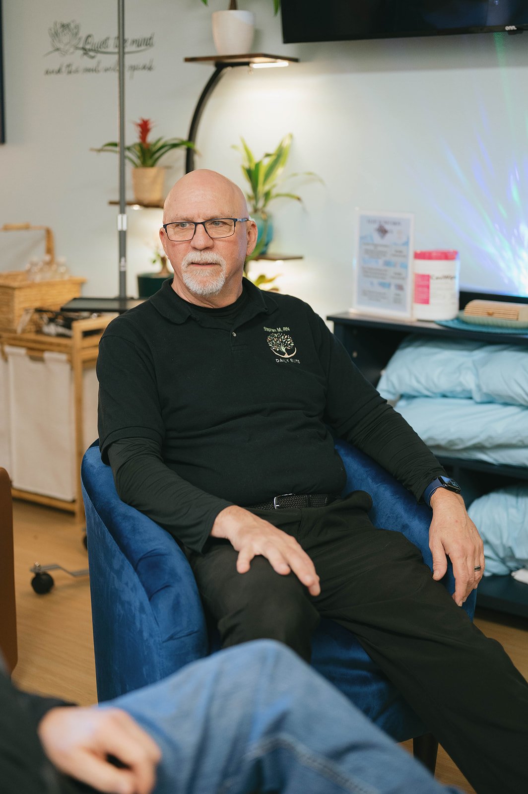 A middle-aged man with glasses and a goatee is sitting in a blue chair in an indoor setting with plants and medical supplies in the background. He is wearing a black shirt with a logo and text.