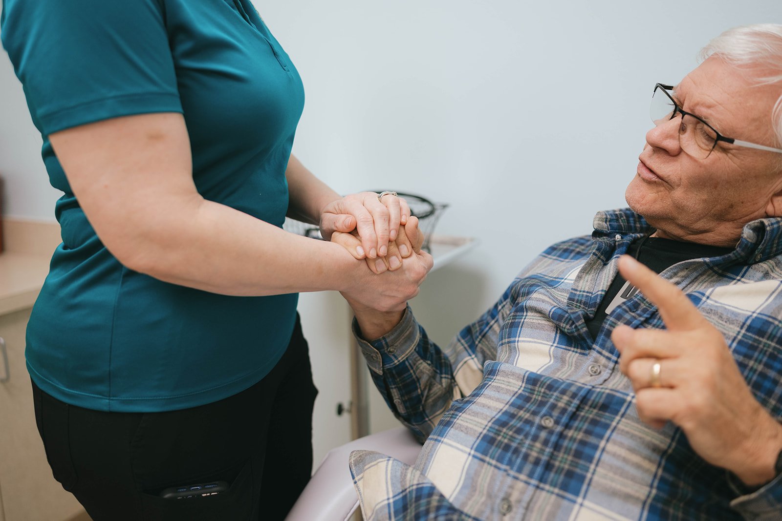 A healthcare worker holding hands with an elderly man seated in a medical office, engaging in conversation.