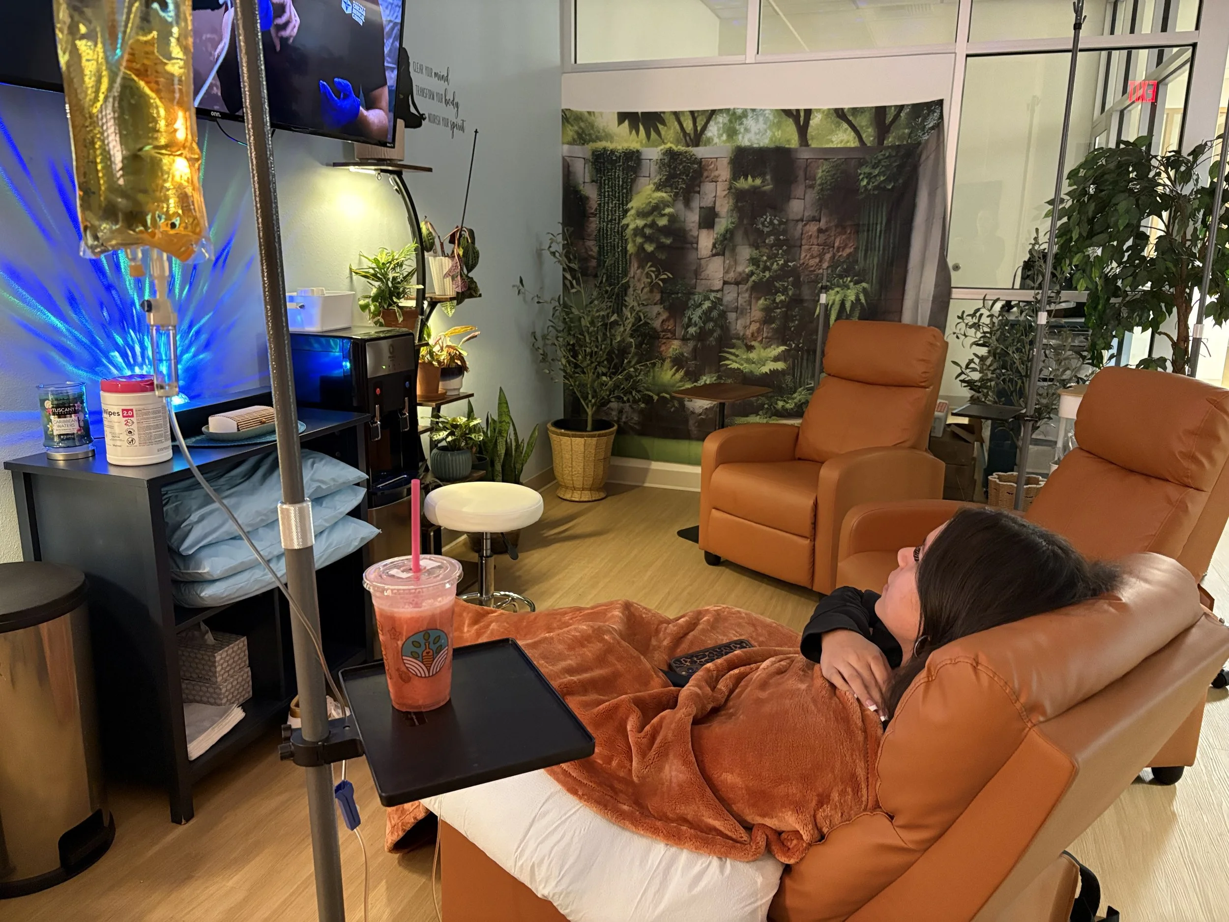 A woman lying on a hospital bed covered with an orange blanket, with an IV pole nearby, in a room decorated with plants and nature-themed wallpaper. There is medical equipment and snacks on a table beside the bed.