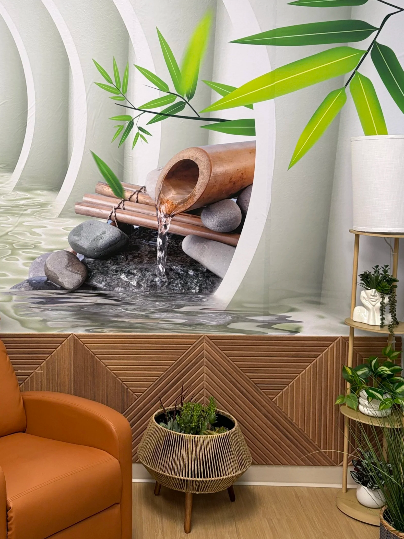 A room with a large tropical-themed mural of bamboo plants and rocks, a tan wooden side table with a potted plant, a cozy orange leather armchair, a woven basket with greenery, and a wooden floor.