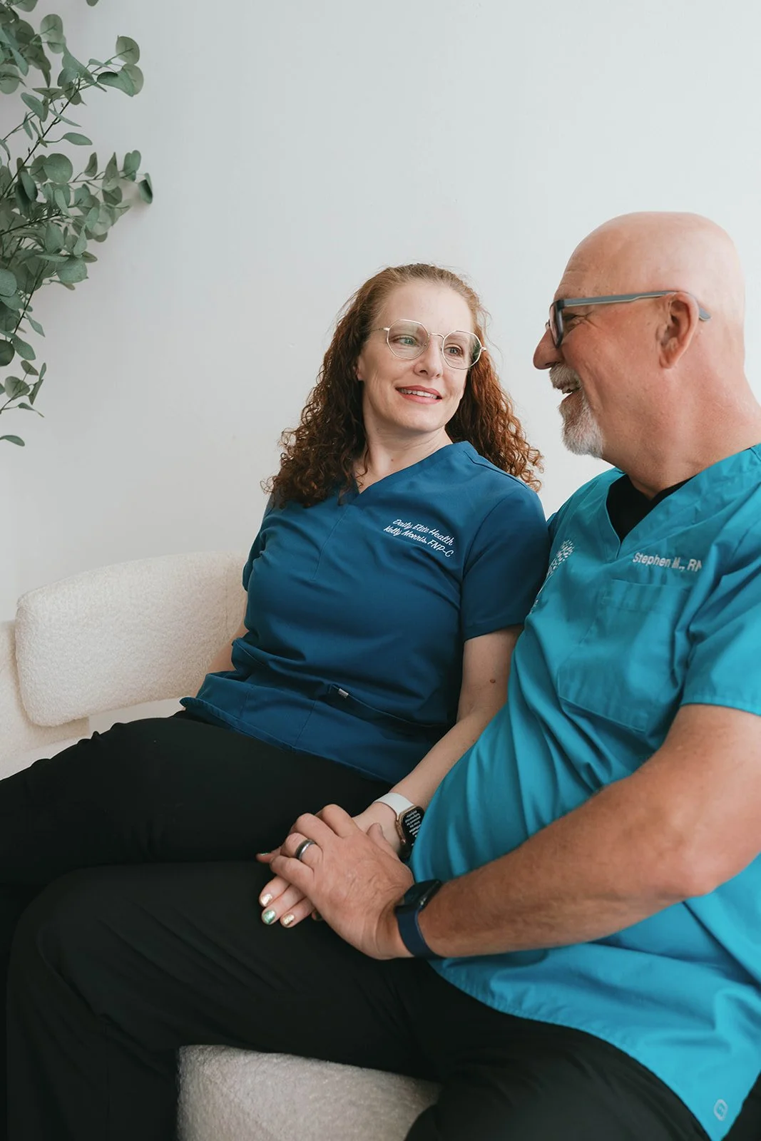 A female healthcare professional with red curly hair and glasses sitting on a beige couch, holding hands with a male healthcare worker with a beard and glasses, both wearing blue scrubs and sharing a moment of connection in a waiting room with a plan
