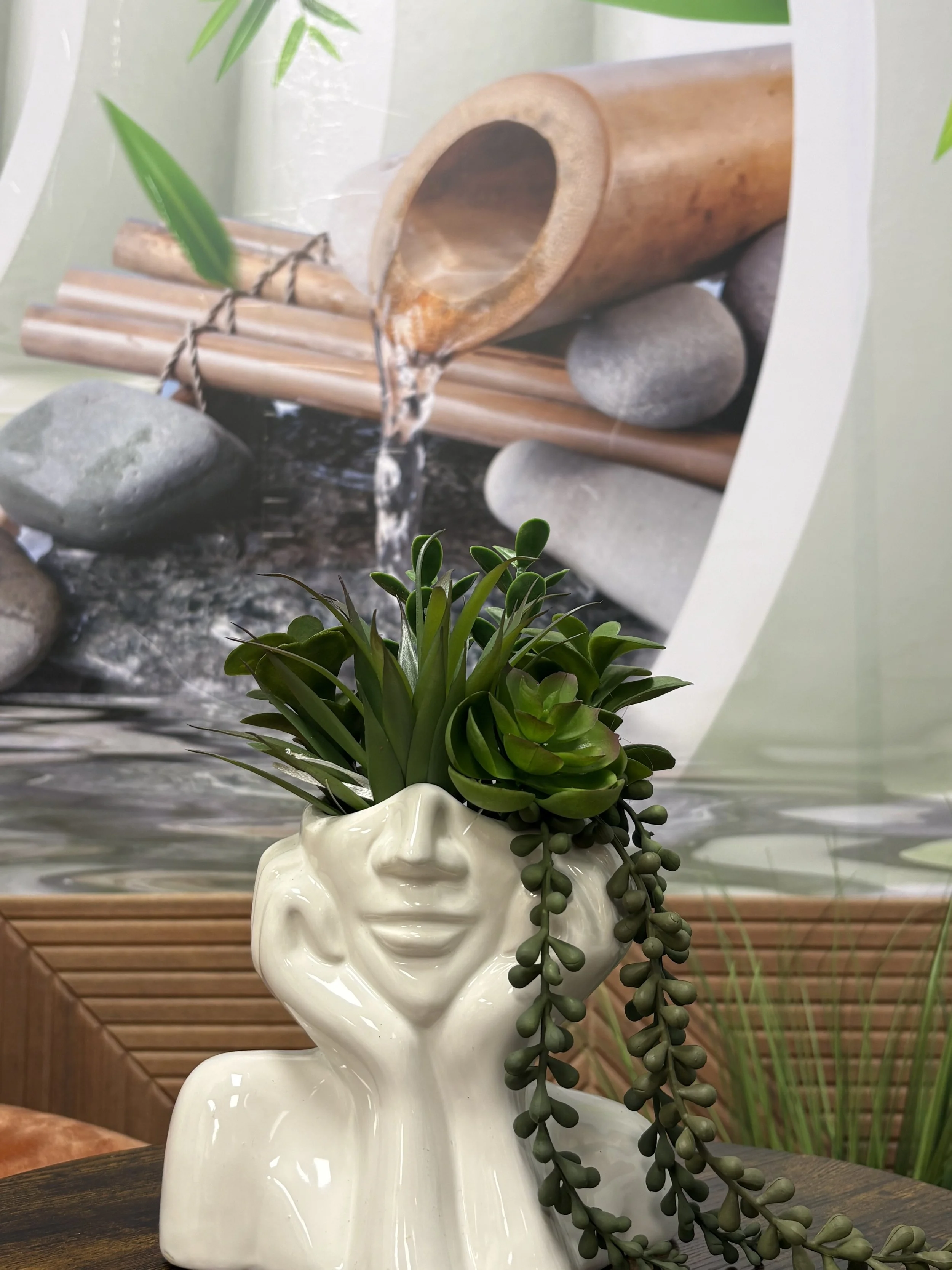 A beige ceramic skull-shaped vase holding a succulent plant with trailing dark green leaves, placed on a wooden surface with a blurred background of wooden panels and indoor plants.