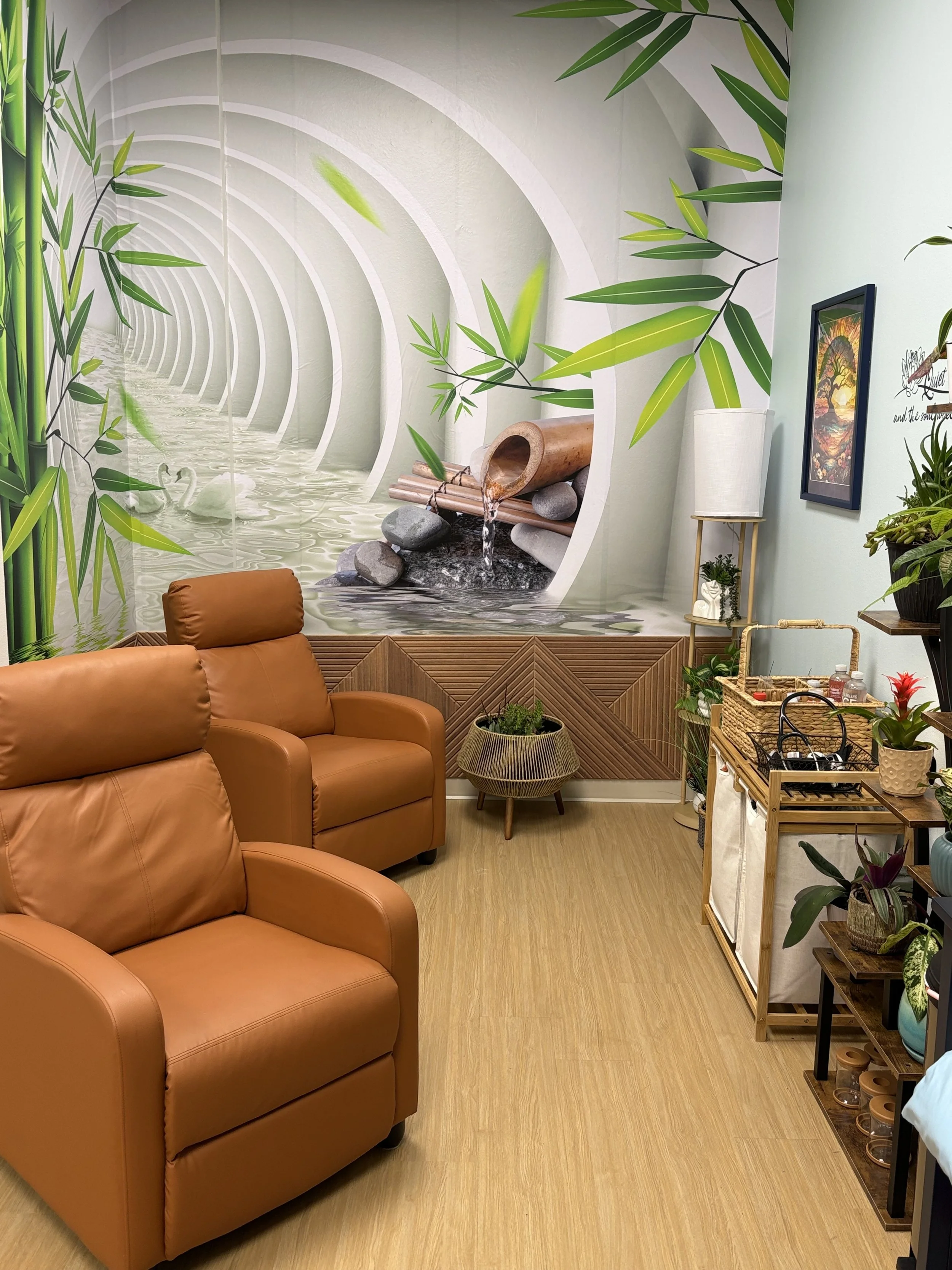 A cozy room with tan leather sofas, a wooden floor, numerous indoor plants, a wicker basket table, and a large mural of bamboo and water landscape on the wall.