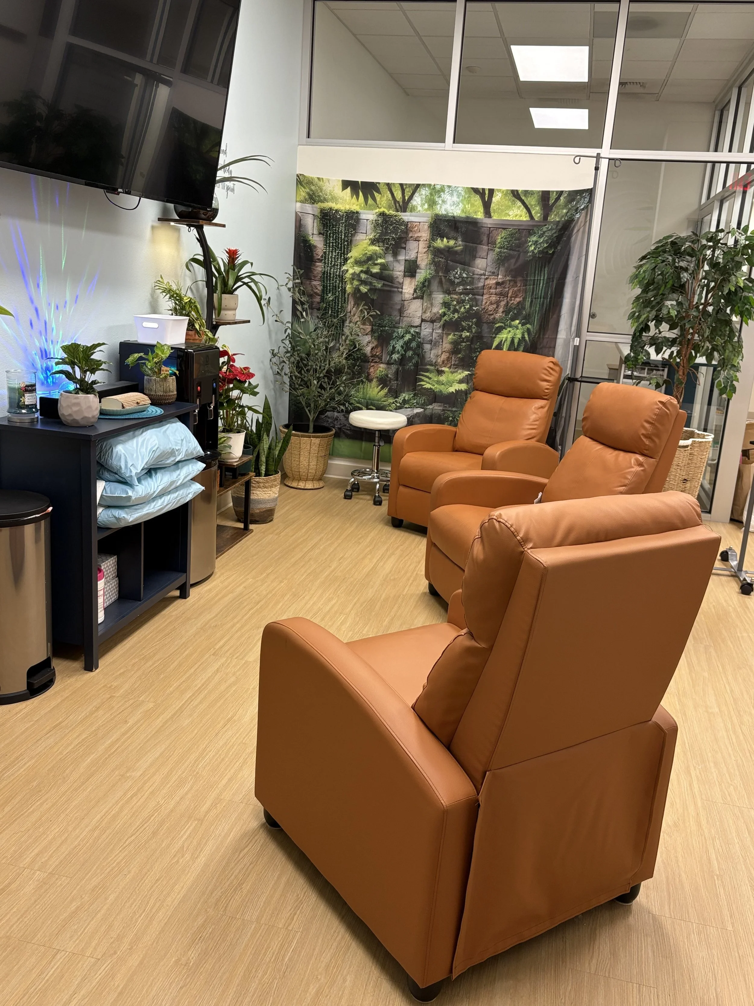 An indoor medical or office waiting area with three tan leather chairs, a small black table with artwork, plants, and decorative items, a wall with a large TV monitor, and a nature-themed backdrop on a glass wall.