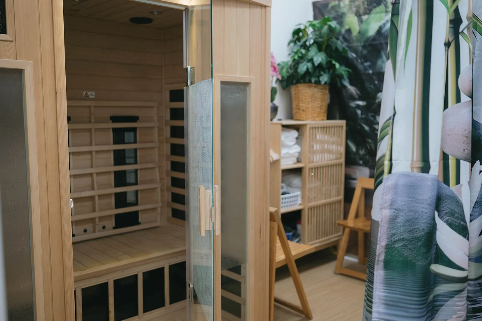 Interior of a room featuring a wooden sauna with a glass door, wooden shelves with folded towels, a wicker basket with a plant, and a curtain with a leaf pattern.