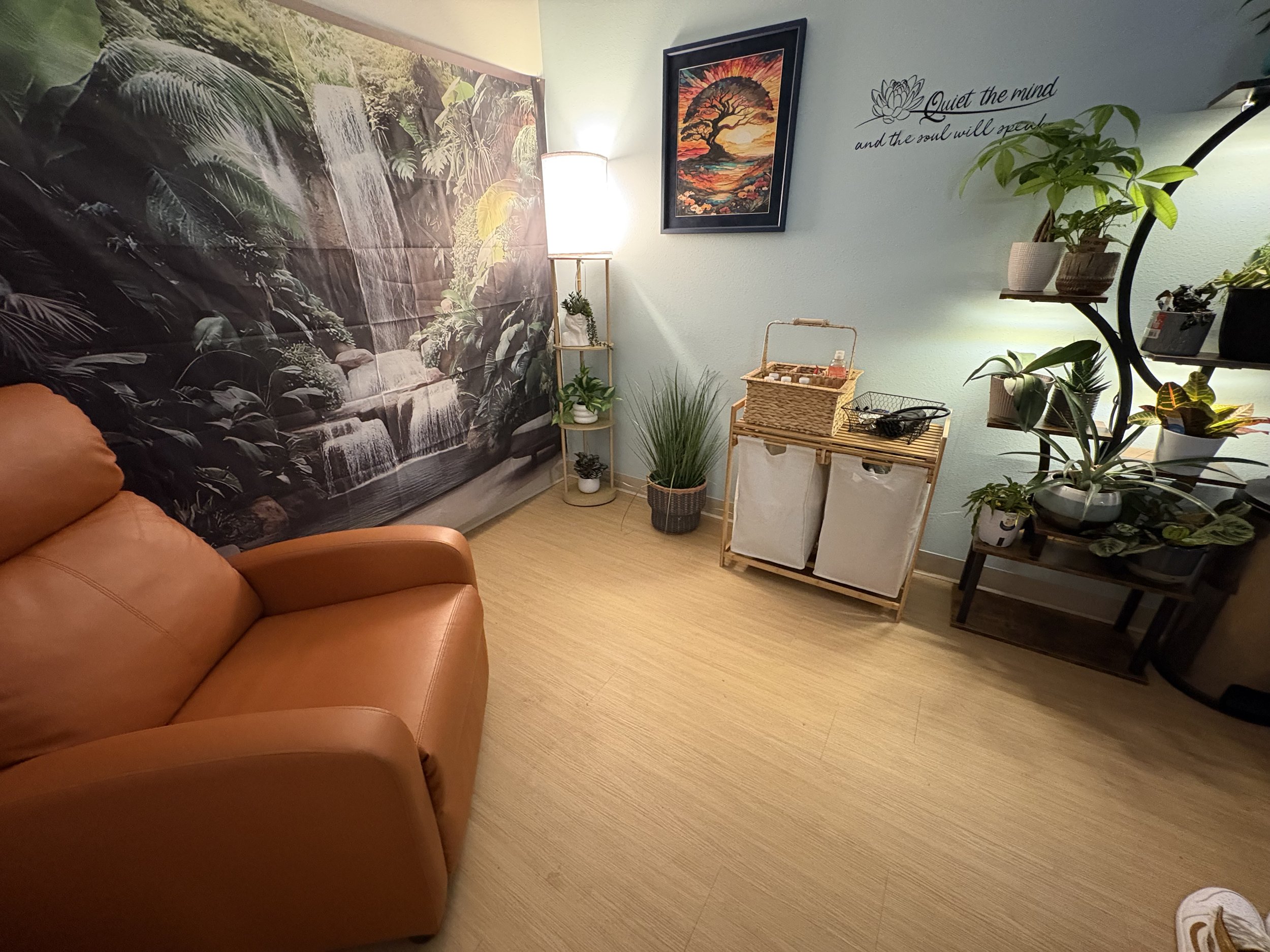 A cozy room with a jungle-themed wall mural featuring dense leaves and a waterfall, a tan leather sofa, a small wooden shelving unit with potted plants, a lamp, a framed sunset tree painting, and indoor plants on shelves and a side table.