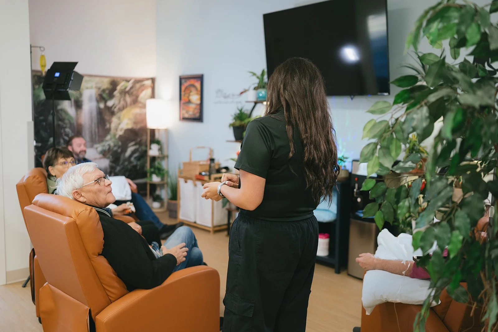 A woman standing and speaking to a seated group of elderly people in a living room decorated with plants, paintings, and a large TV.