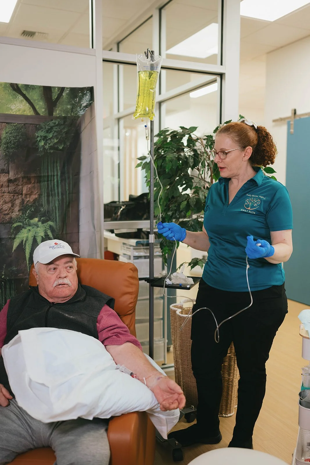 A woman nurses an elderly man with an IV drip attached to his arm in a healthcare setting.