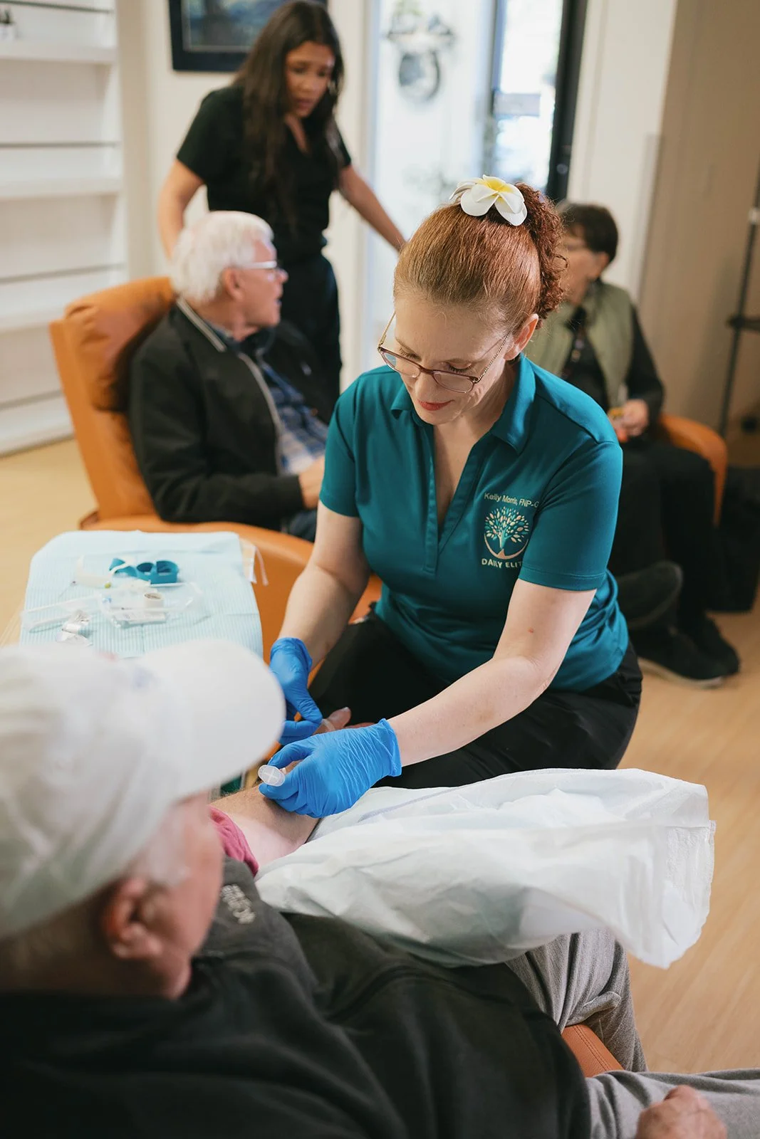 Healthcare professional in teal uniform administering a medical injection to an elderly patient lying on a bed, while two other elderly patients sit nearby in a clinic or medical setting.