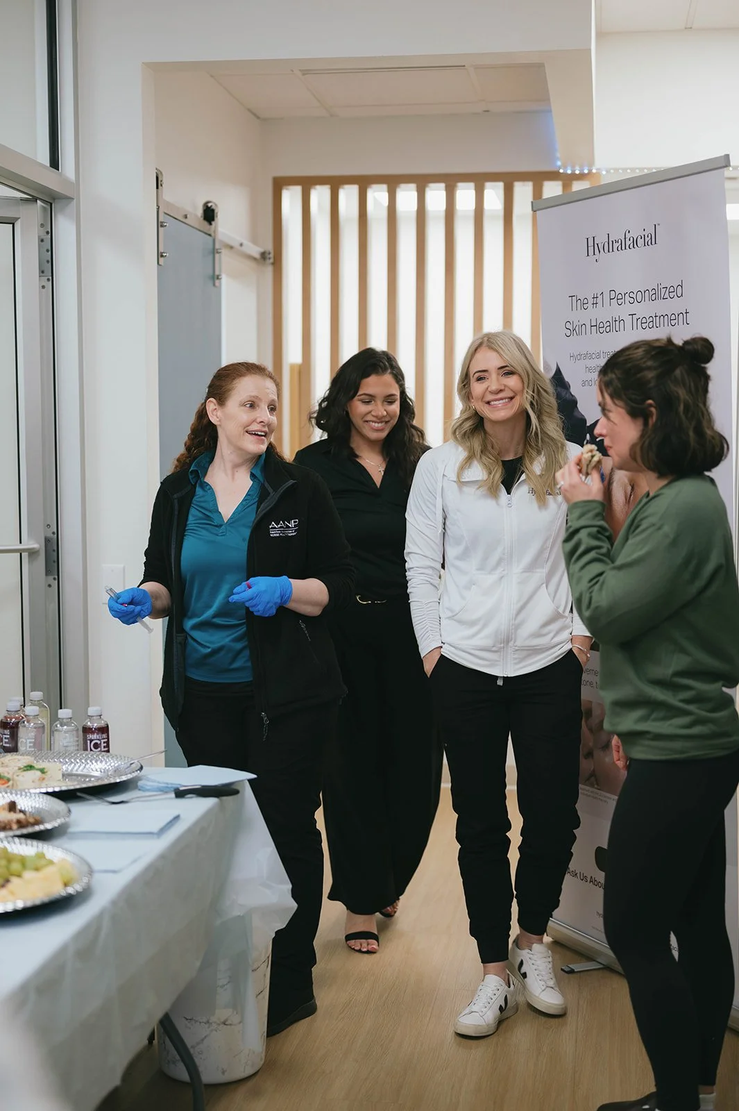 Group of four women standing indoors at a health event. One woman eating while others chat, with a table of snacks and drinks nearby. Banner promotes personalized skin health treatments.