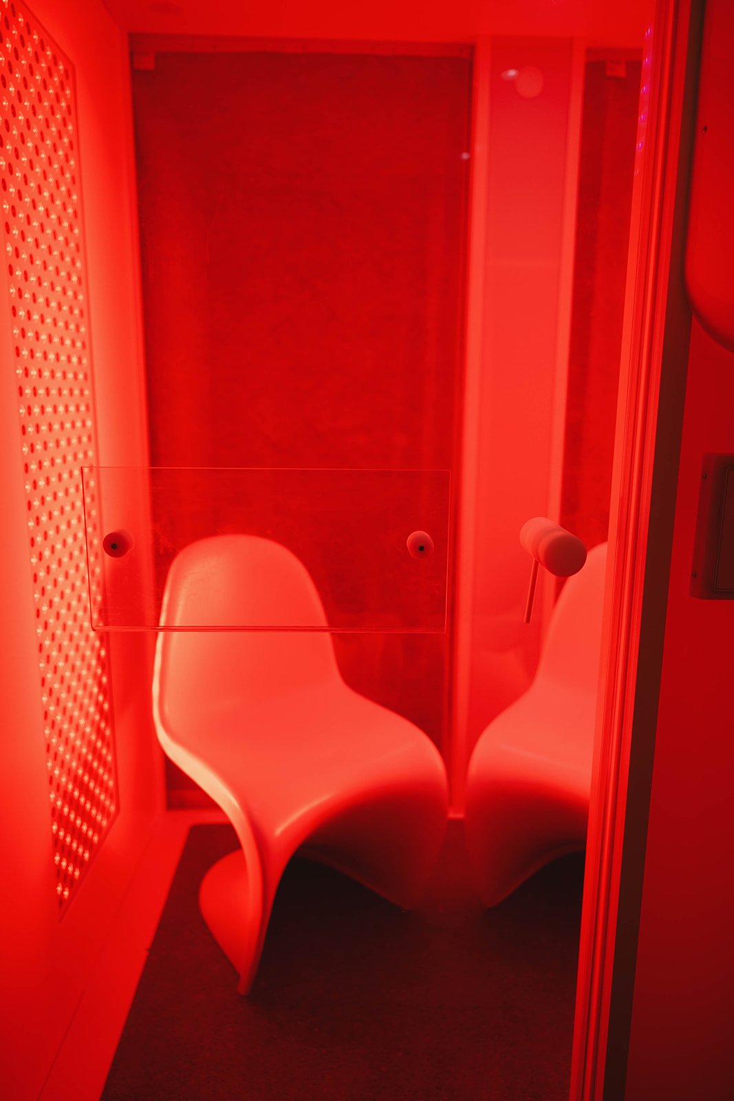 Red-lit pod with a white, modern chair and a glass partition inside a photo booth or lounge area.