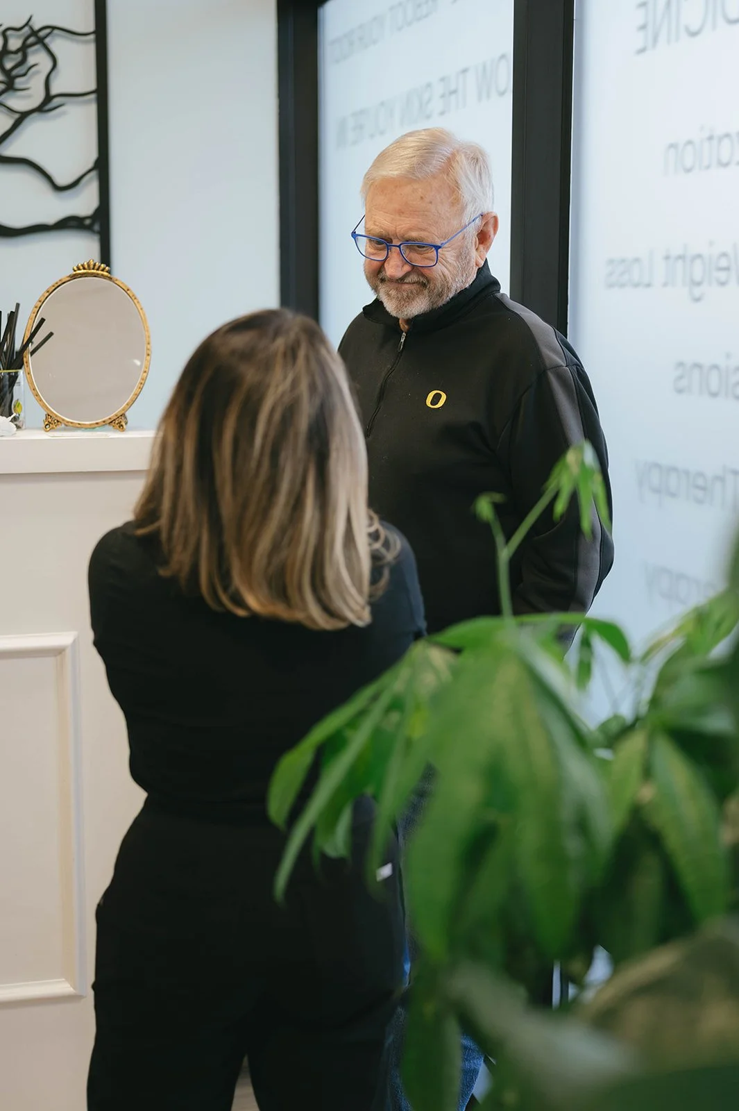A man with glasses and a gray beard wearing a black jacket talking to a woman with shoulder-length brown hair in an office setting.