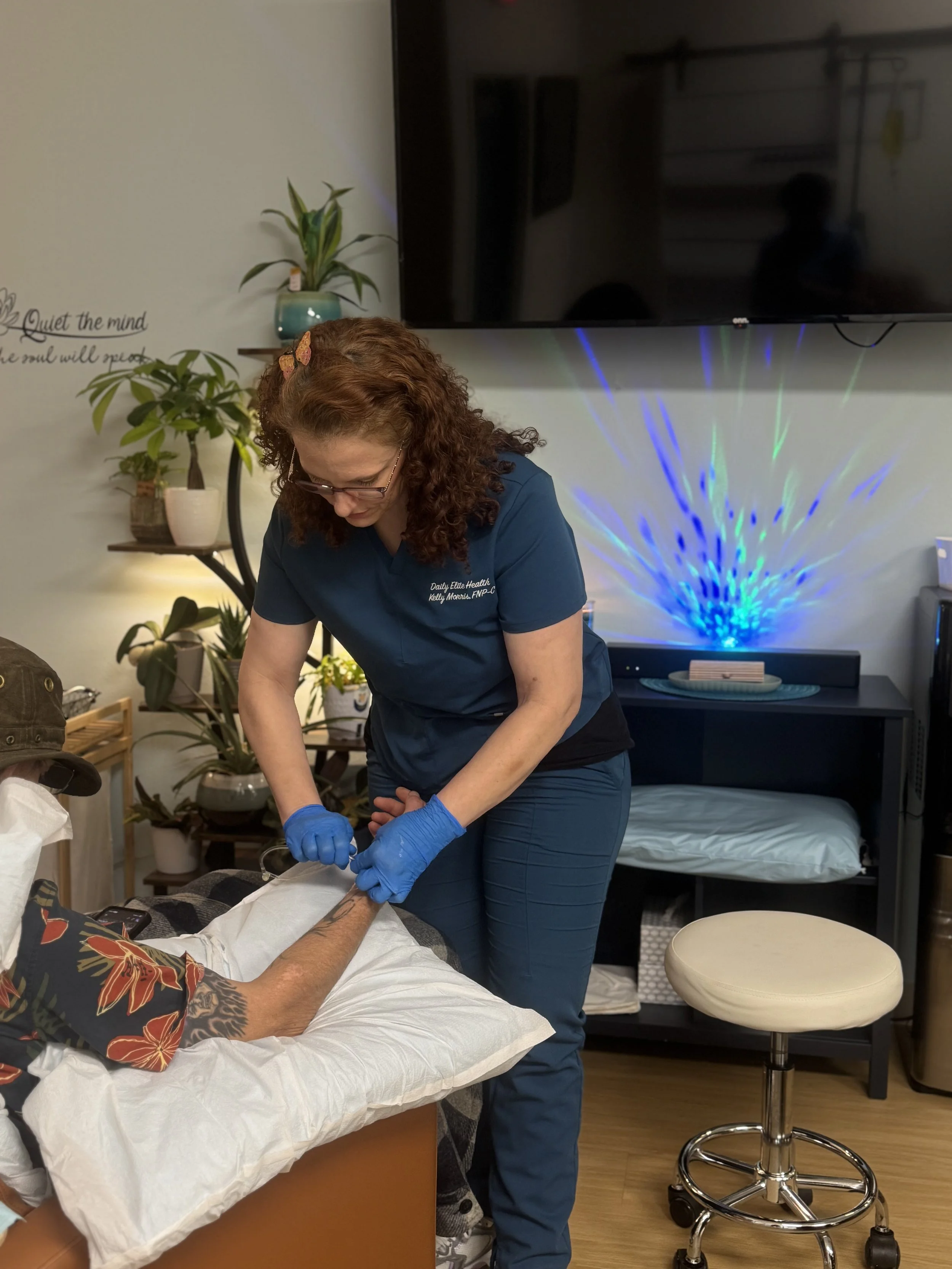 A healthcare professional wearing scrubs and gloves provides medical care to a patient lying on a bed in a treatment room. The room has plants, a large television, and decorative lighting.