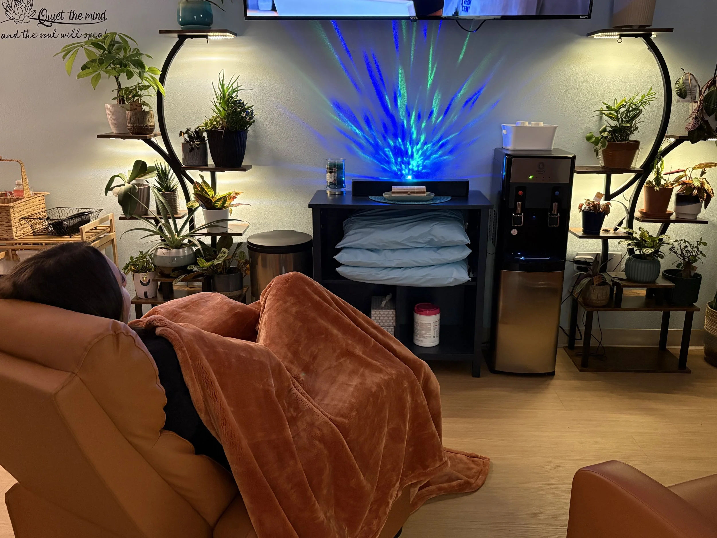 Living room with a tan couch covered with an orange blanket, a black shelf with stacked pillows, and multiple potted plants on shelves and the floor. A wall-mounted TV displays colorful lights, and there's a quote on the wall: 'Quiet the mind and the