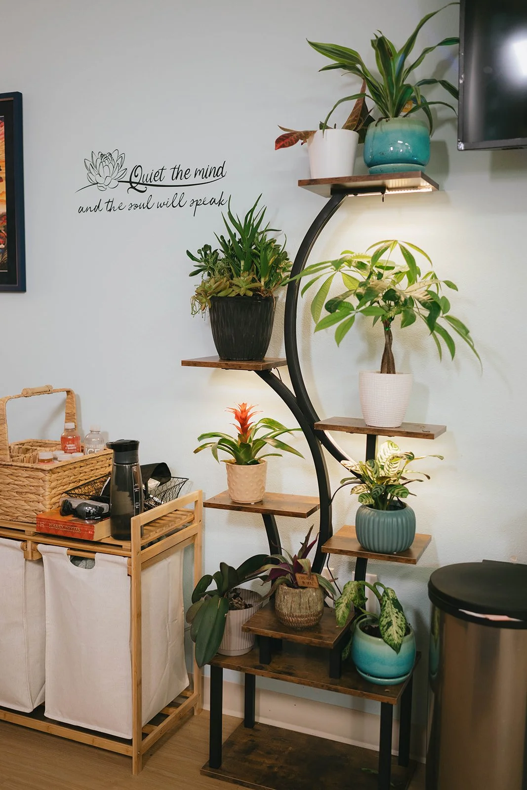 A corner of a room with a curved wooden shelf holding various potted plants. A wall decal reads, 'Quiet the mind and the soul will speak,' next to a framed picture. A small wooden cart with bottles, a thermos, and other items is nearby, and a black t