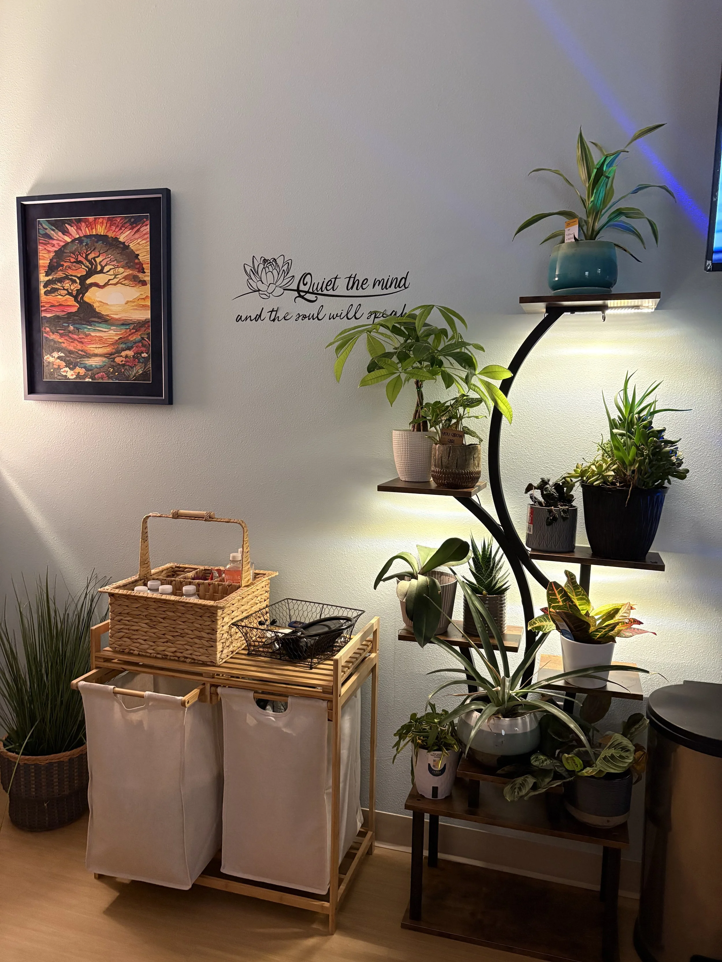 Indoor scene with a variety of potted plants on shelves and a table, a framed colorful painting of a tree on the wall, and a quote on the wall that reads 'Quiet the mind and the soul will speak.'