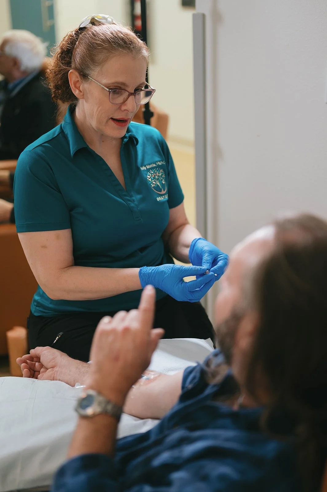 A healthcare worker wearing glasses, a teal polo shirt with a logo, and blue gloves, prepares to give a vaccination to a patient lying in a hospital bed. The patient, a man with long hair and a beard, is sitting up and gesturing with his hand.