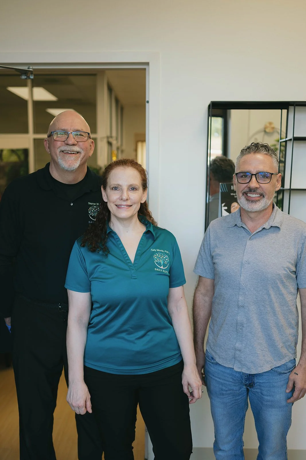 Three people standing together indoors smiling. The woman in the middle wears a teal shirt with logo, the man on the right wears gray, and the man on the left wears black. There is a mirror behind and a window in the background.