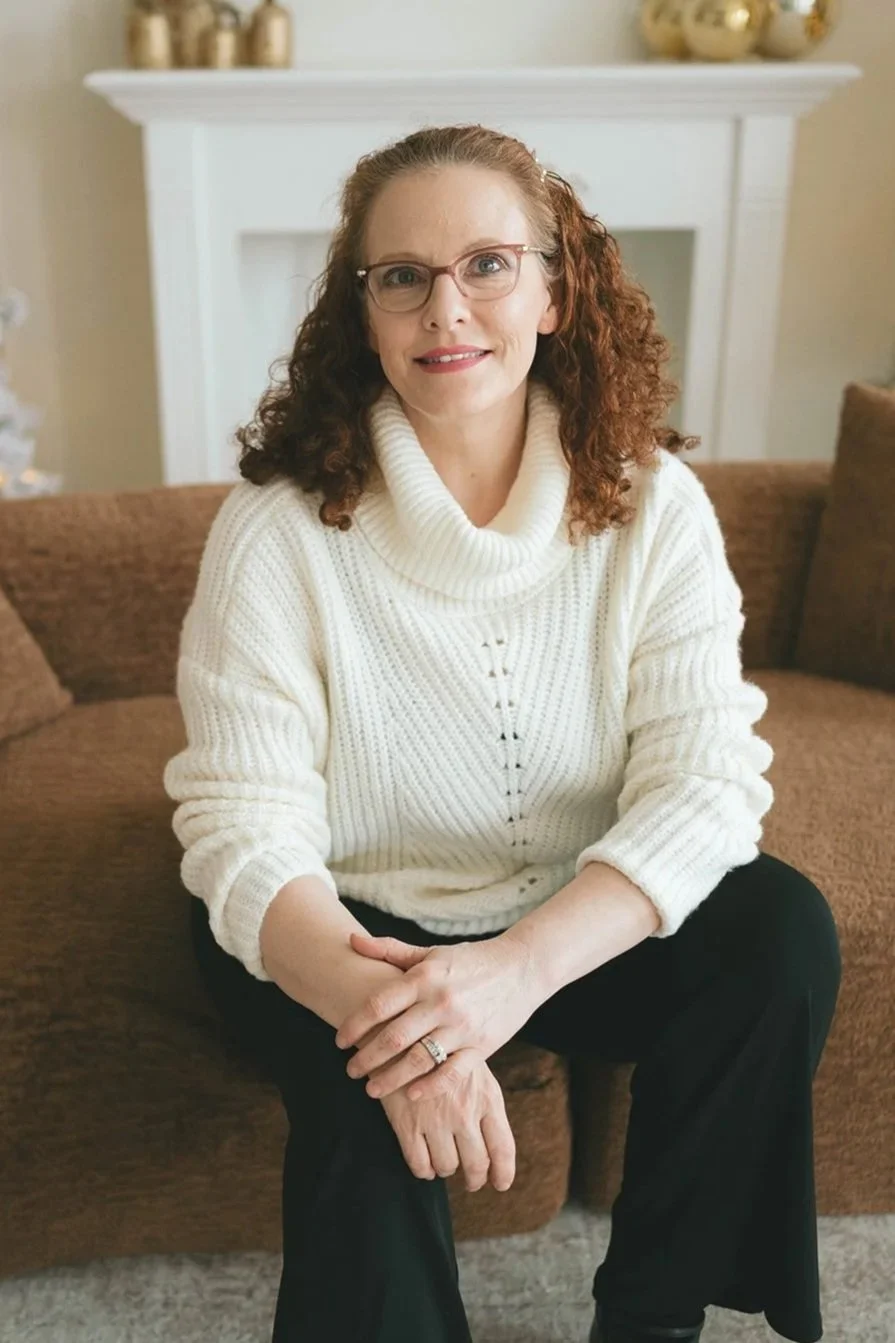 A woman with curly red hair, wearing glasses and a white turtleneck sweater, sitting on a brown couch in a cozy living room with a fireplace decorated with gold ornaments in the background.
