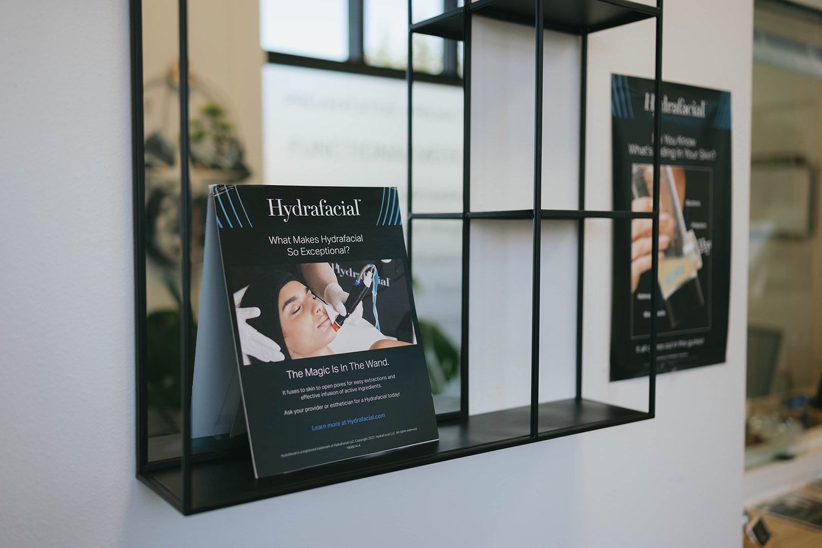 Brochure on hydrafacial treatment on a black wire shelf, showing a woman receiving a hydrafacial in a spa or clinic setting.