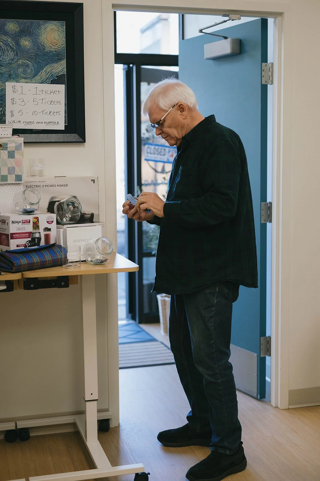 An elderly man with white hair and glasses looking at his phone inside a shop near a display of kitchen appliances and gifts.