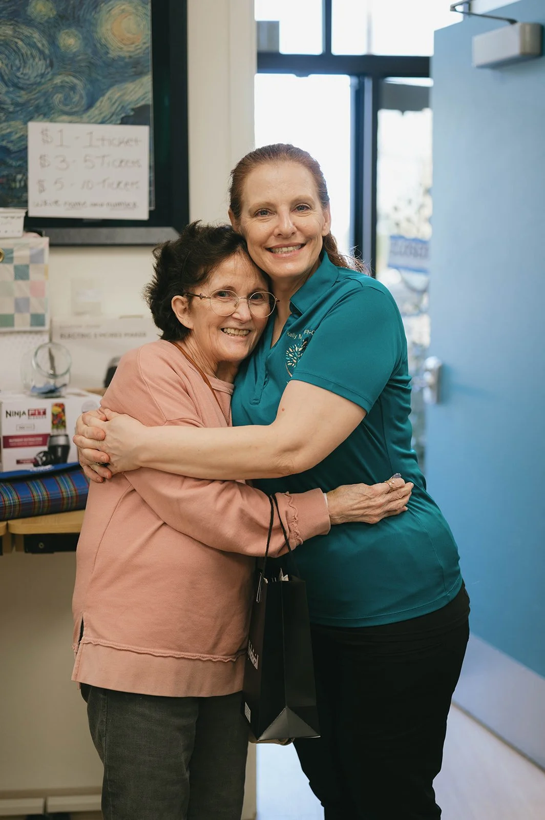 Two women embracing inside a room, smiling at the camera. One woman is wearing a pink top and glasses, and the other is in a teal shirt with a logo.