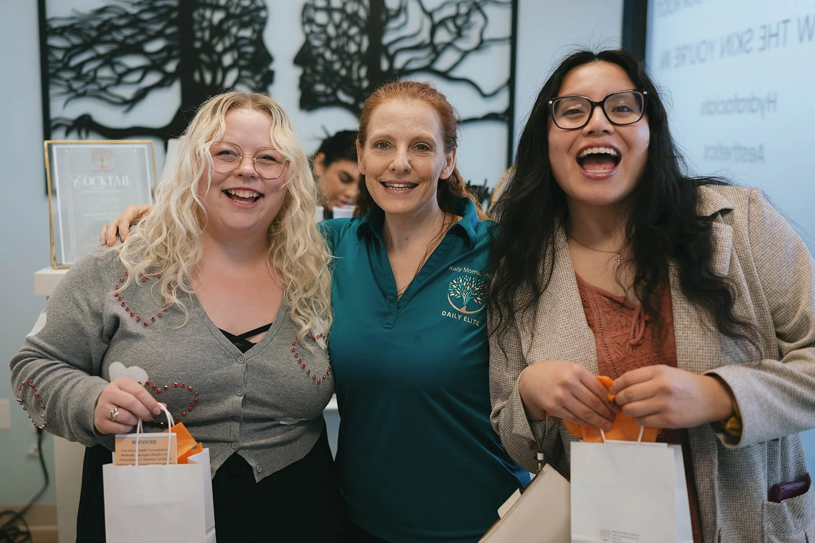 Three women smiling and posing together indoors, holding shopping bags, with a large wall art piece and a screen in the background.