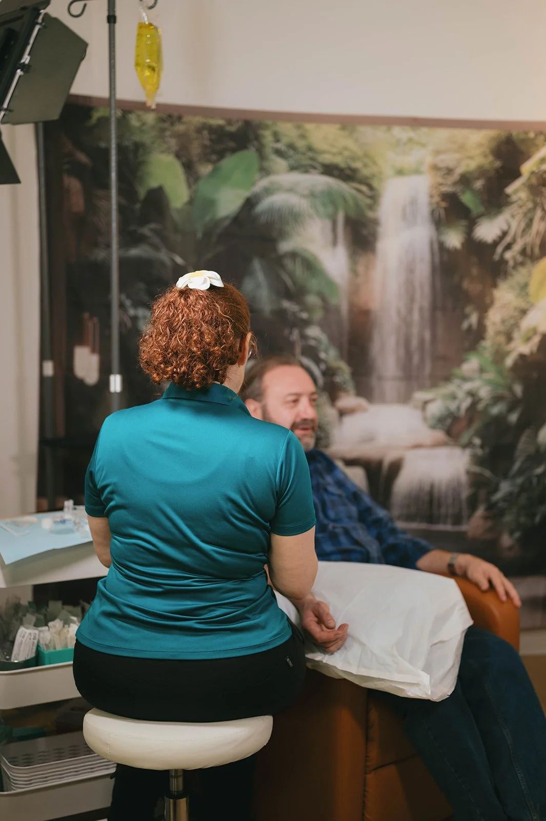 A woman with curly red hair, wearing a teal shirt, sits on a white stool facing a man with a beard, wearing a blue checkered shirt, who is seated in a brown armchair. A waterfall mural with lush greenery decorates the background, and a medical IV dri