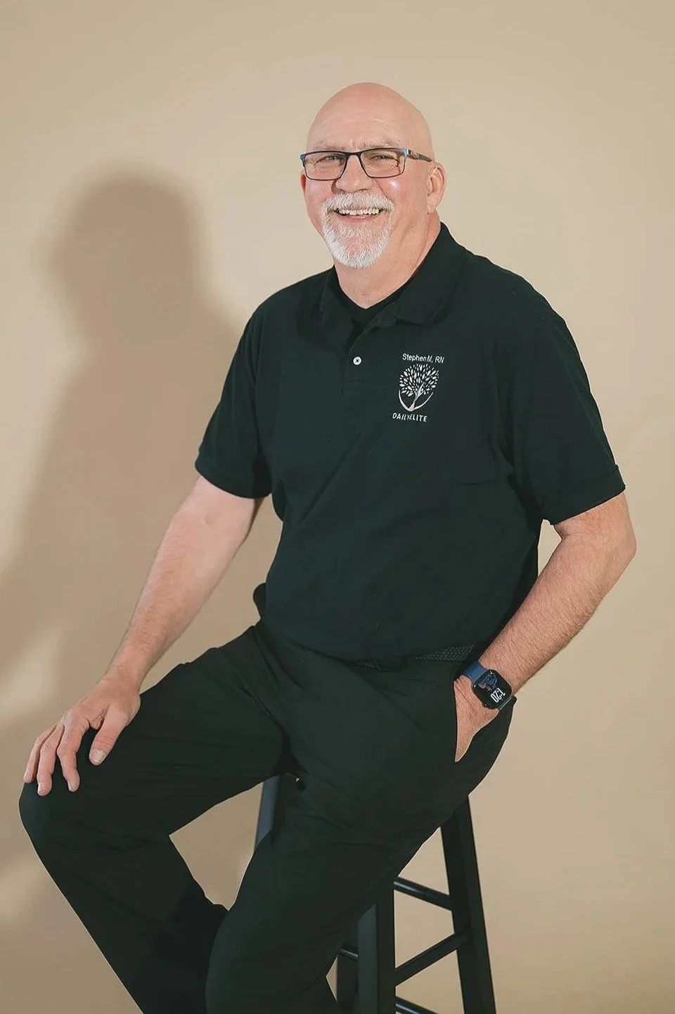 A middle-aged man with glasses, a white beard, and a bald head, smiling and wearing a black polo shirt with a tree logo and the text "Stephen M, RN" and "DAILY ELITE". He is sitting on a stool against a beige wall.
