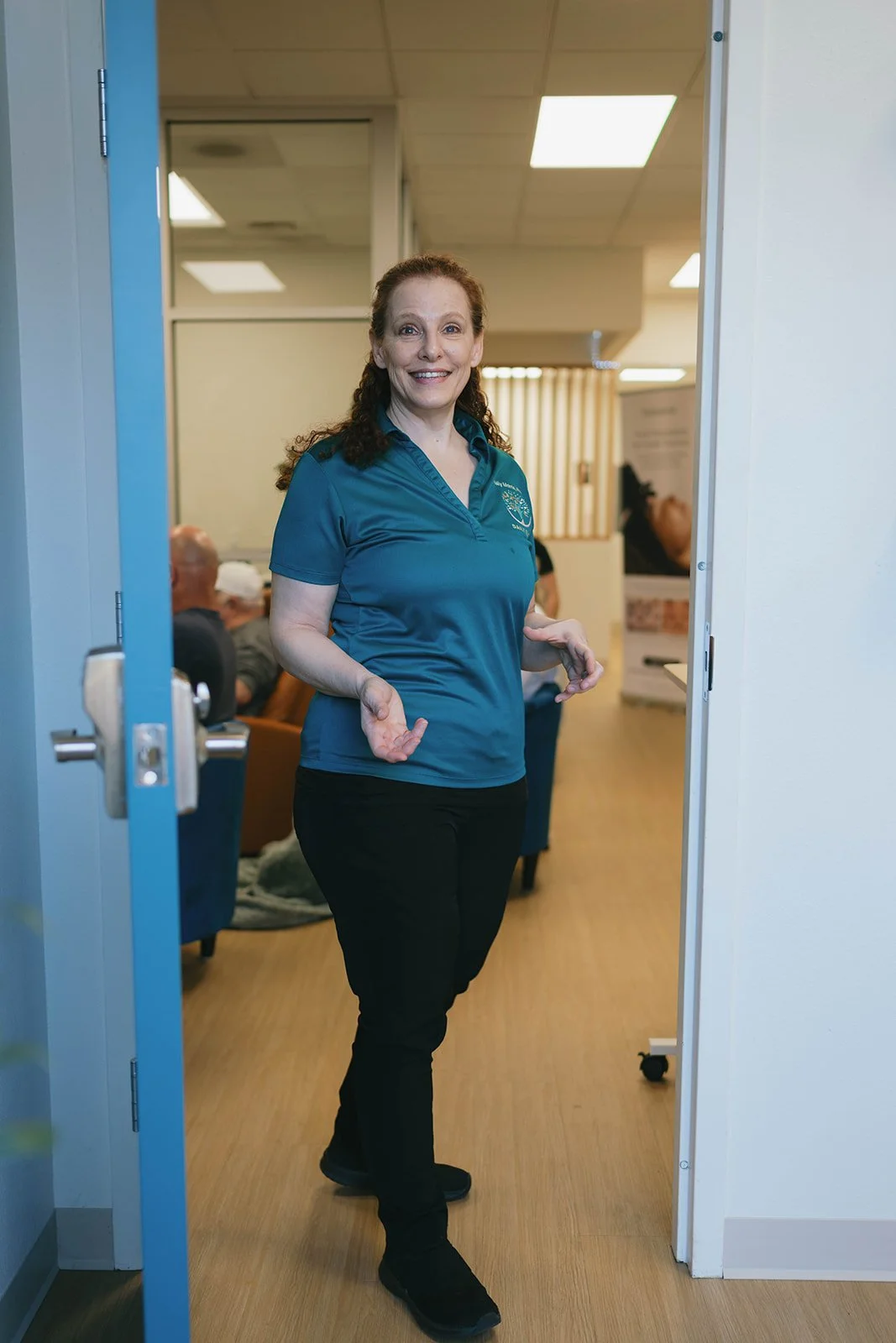 A woman with red hair and a blue polo shirt standing in a doorway, smiling, with people seated in the background.