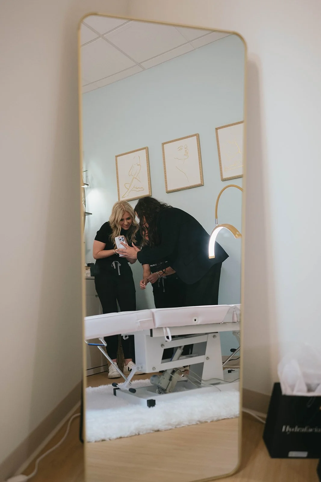Two women looking at a phone, seen through a mirror in a room with medical or spa equipment and art on the wall.