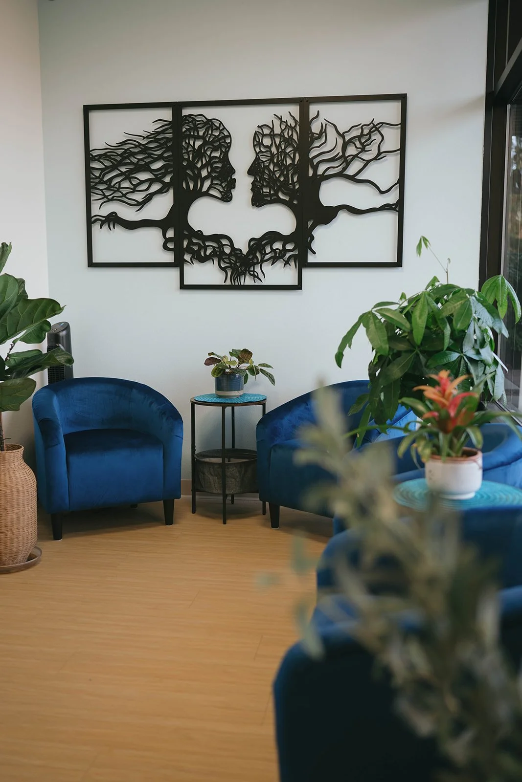 Interior of a living room with two blue velvet chairs, a small black side table with a potted plant, and a large wall art piece depicting two face profiles made of tree branches. Various green plants are also present.