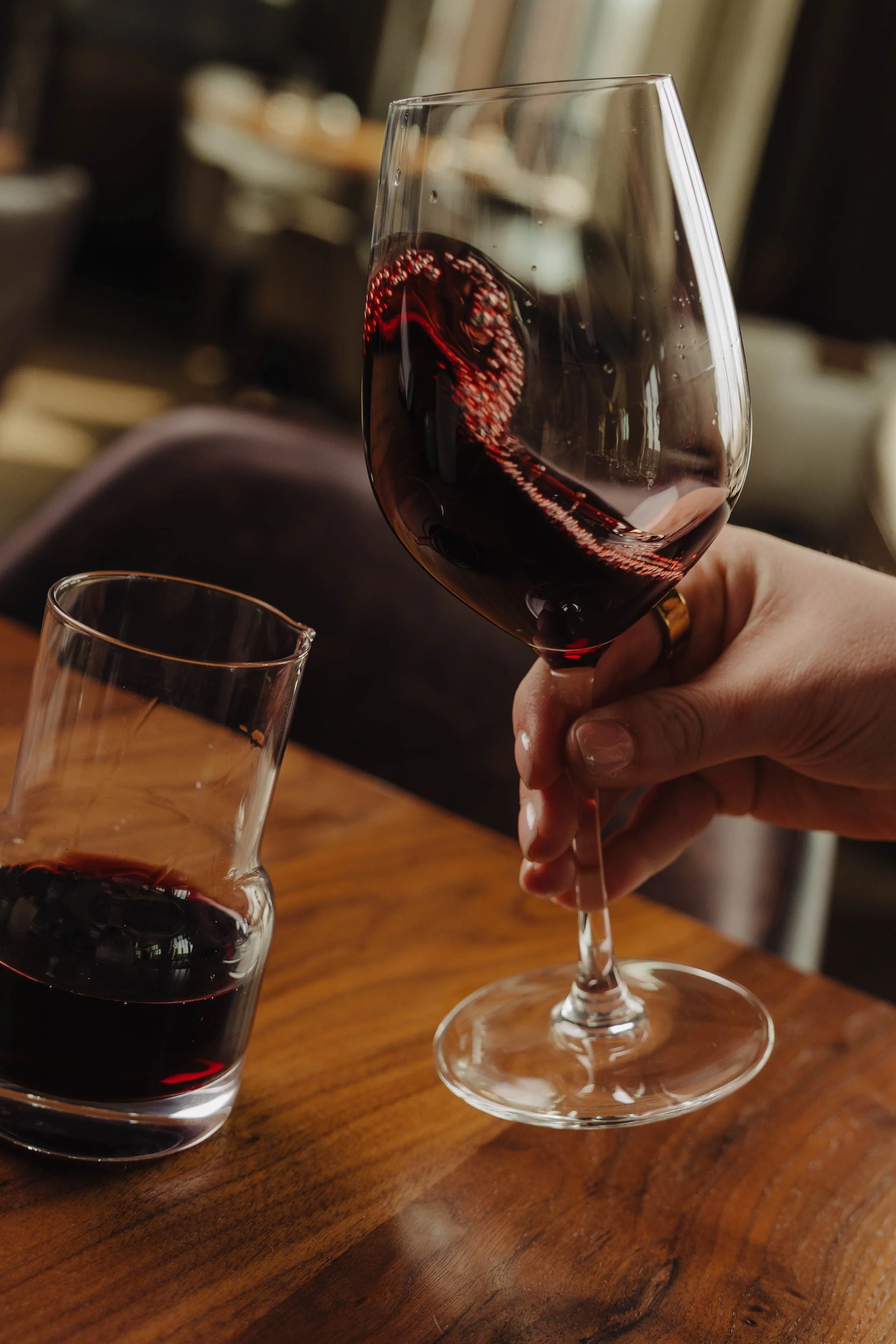A hand holding a glass of red wine next to a wine decanter on a wooden table.