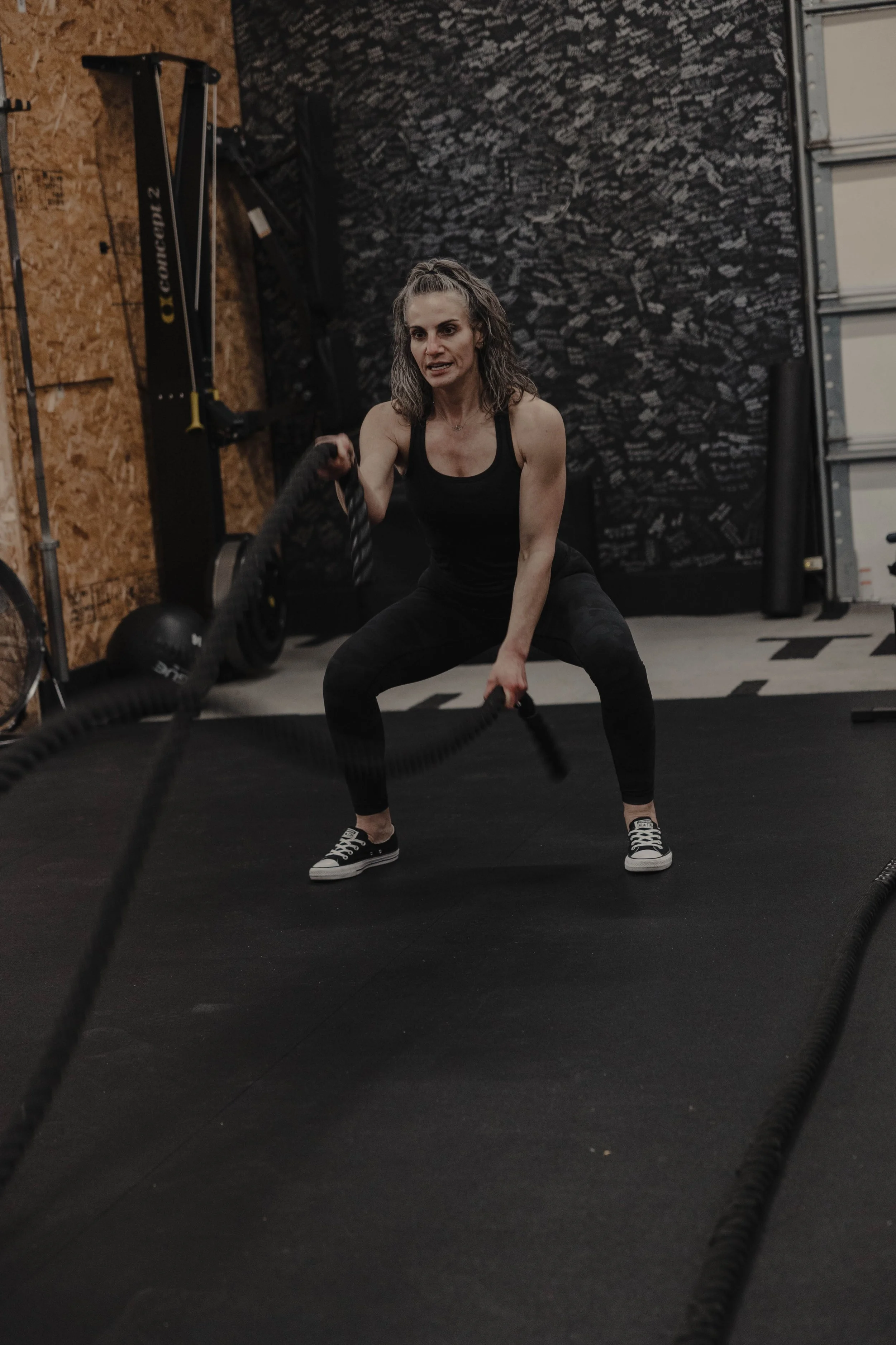A woman exercising with battle ropes in a gym, wearing a black tank top, black leggings, and sneakers. The background includes gym equipment and a wall with writing.