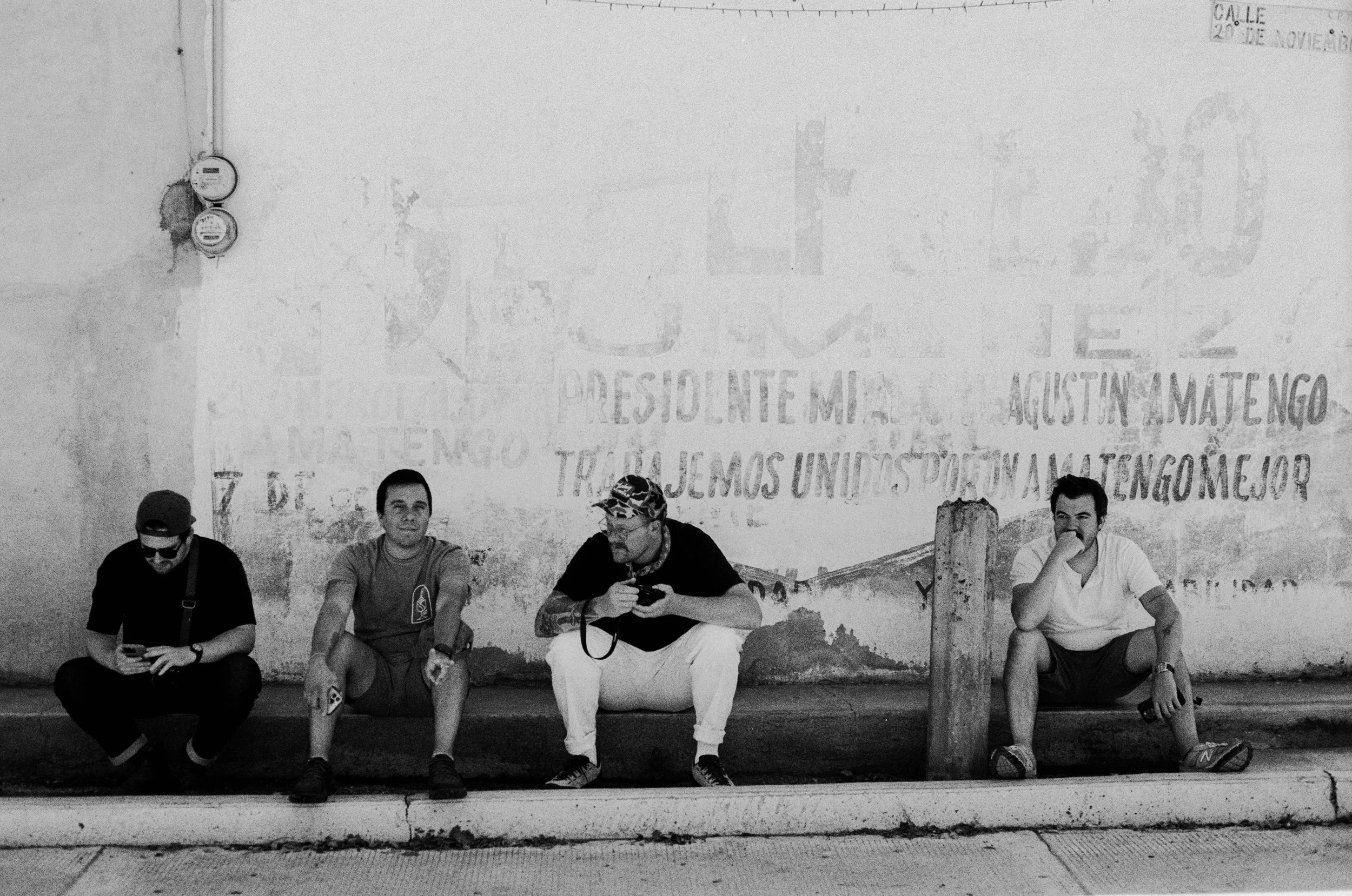 Four men sitting on a sidewalk against a wall with faded graffiti.